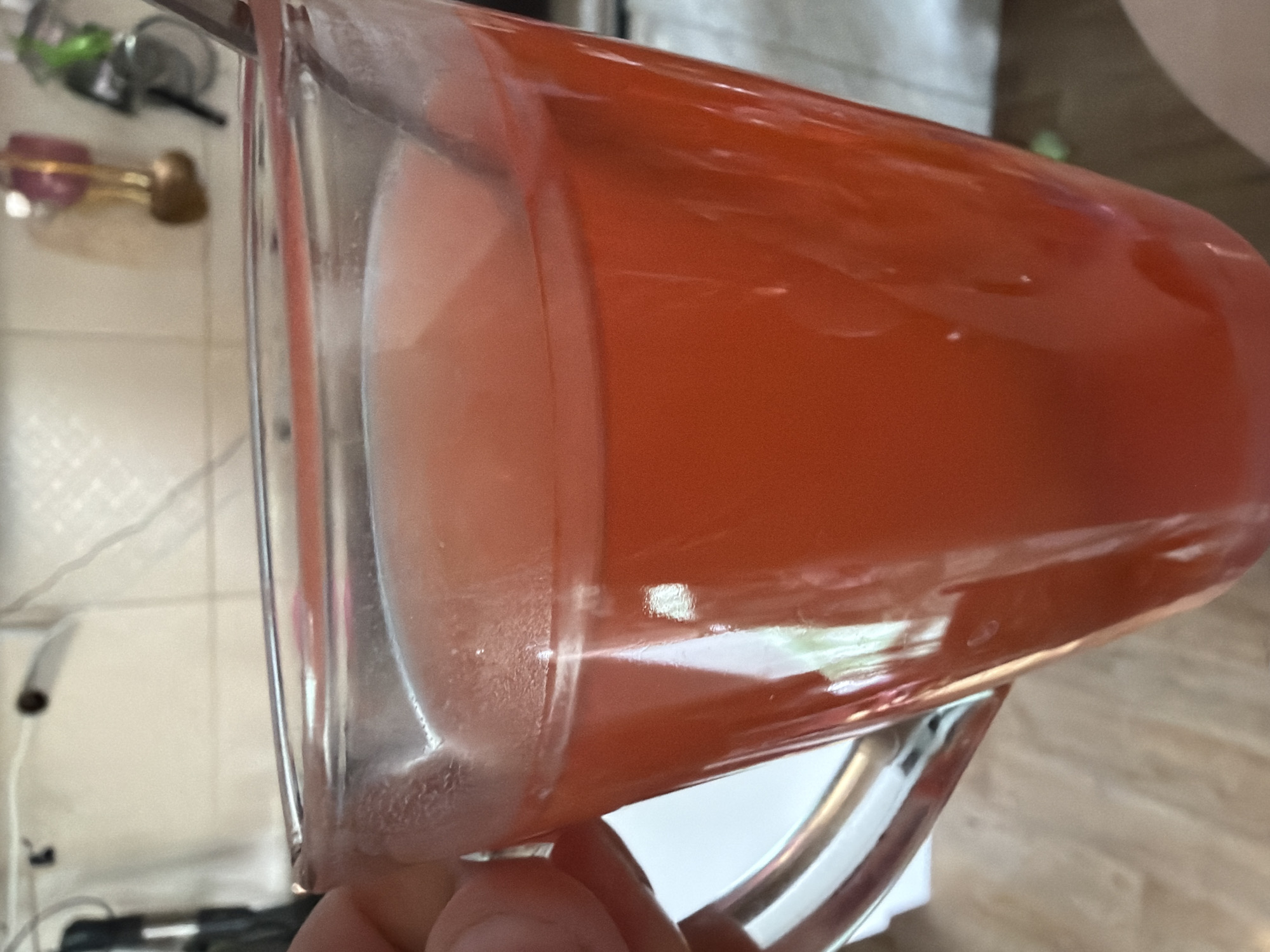 The image is a close-up, eye-level shot of a clear glass mug filled with a red-colored liquid, likely a beverage such as juice or tea. A spoon is partially submerged in the liquid, suggesting it has been stirred or is being used to drink. The mug is held by a hand, only partially visible on the left side of the frame, with fingers gripping the handle. The background is out of focus but appears to be an indoor setting with what looks like a kitchen counter and some hanging objects. The lighting is bright, creating reflections on the glass mug. The overall scene is simple and domestic, with no identifiable landmarks or specific urban features that would allow for pinpointing a location within Lopatino, Russia.