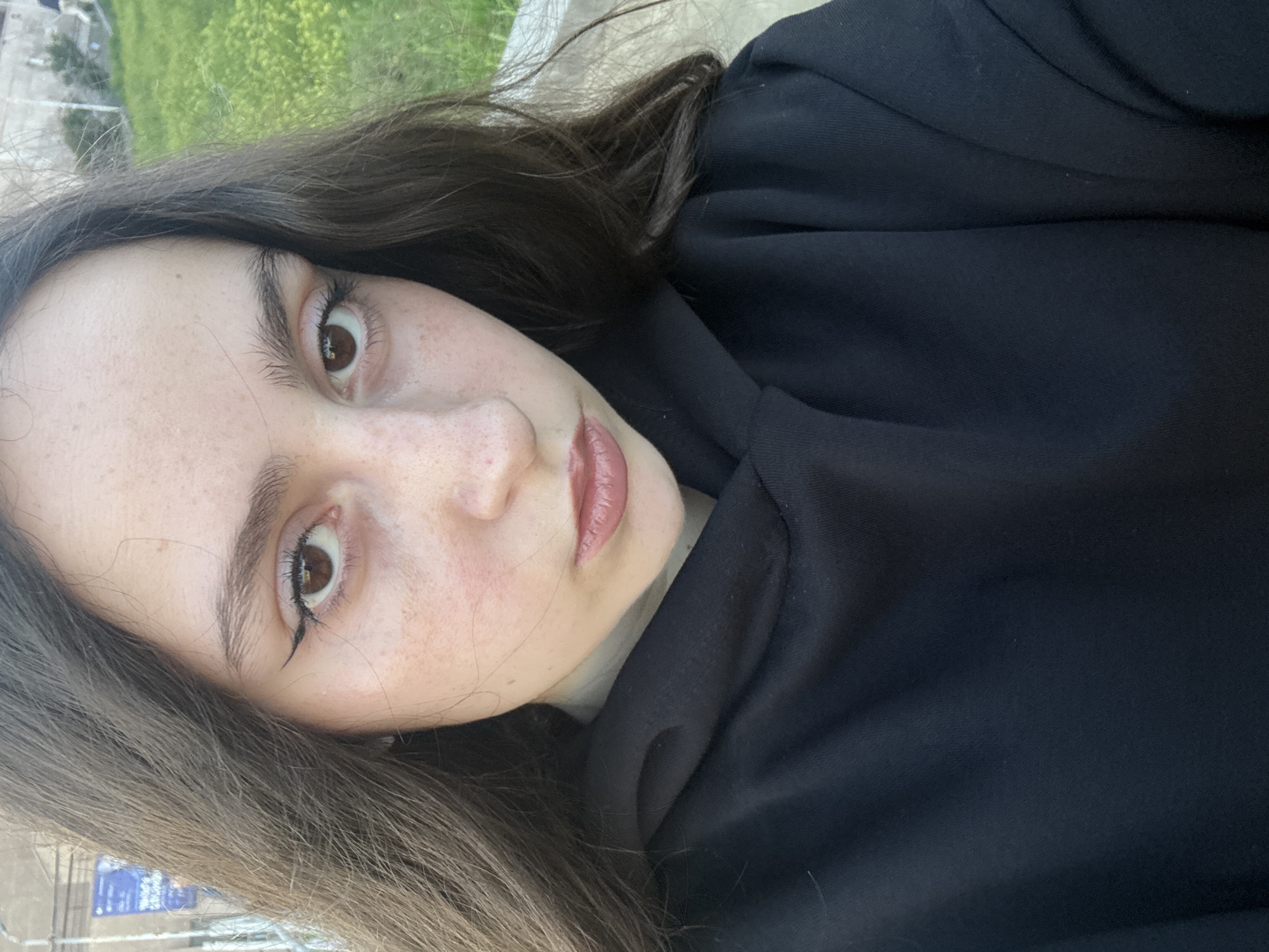 A close-up, slightly tilted portrait captures a young woman outdoors in Yerevan, Armenia. The woman, positioned diagonally in the frame, has long, dark brown hair, fair skin with noticeable freckles, and brown eyes accentuated with black eyeliner. Her lips are painted a glossy, rosy-nude shade, and she maintains a neutral, direct gaze towards the camera. She is wearing a black hoodie or sweatshirt.
The background is softly blurred, indicating a shallow depth of field, which keeps the woman as the primary focus. Above and behind her (due to the tilt) is a vibrant expanse of green foliage, suggesting a grassy area or shrubbery, typical of an outdoor park or garden setting. In the lower left of the image, blurry elements include what appear to be parts of urban infrastructure, possibly a road or sidewalk, and faint blue signs with indecipherable white text. The lighting suggests it is daytime with natural light. The overall impression is a casual, direct portrait taken in an urban green space.