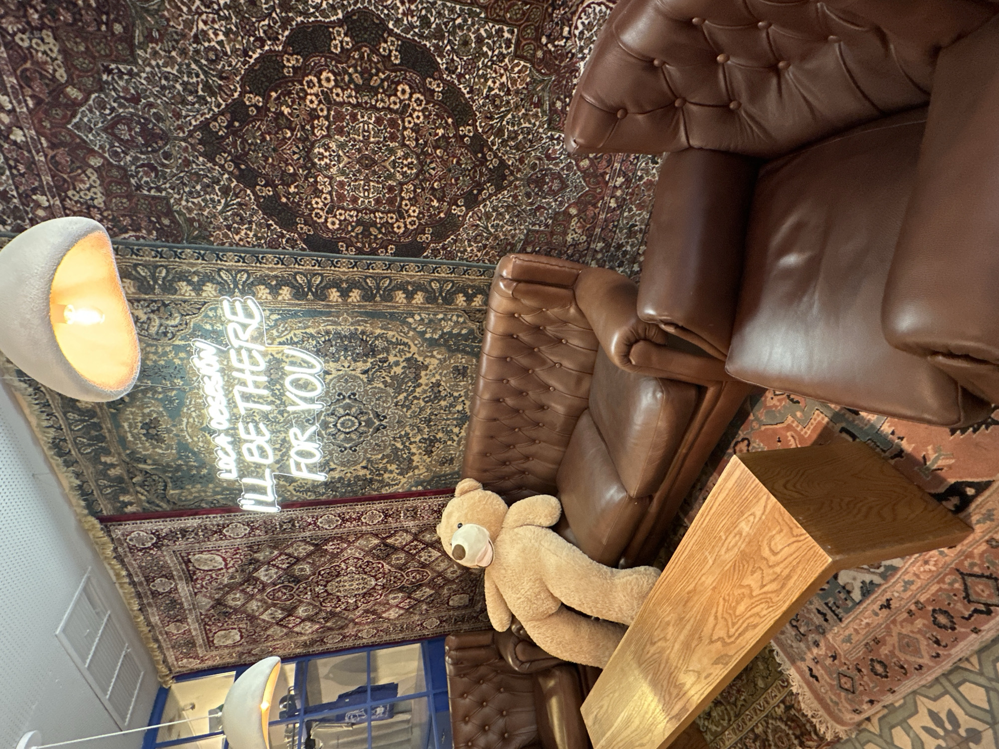 This image depicts an interior scene, likely a cafe or lounge area, with a prominent display of Persian rugs on the wall. In the foreground, a large, plush teddy bear is seated on a brown leather Chesterfield sofa. To the left of the sofa, a wooden coffee table is positioned. The background features more ornate Persian rugs, and a neon sign with the text "LOVA DESSERT I'LL BE THERE FOR YOU" is visible. Two light fixtures hang from the ceiling, casting a warm glow on the scene. The overall ambiance suggests a cozy and eclectic space.