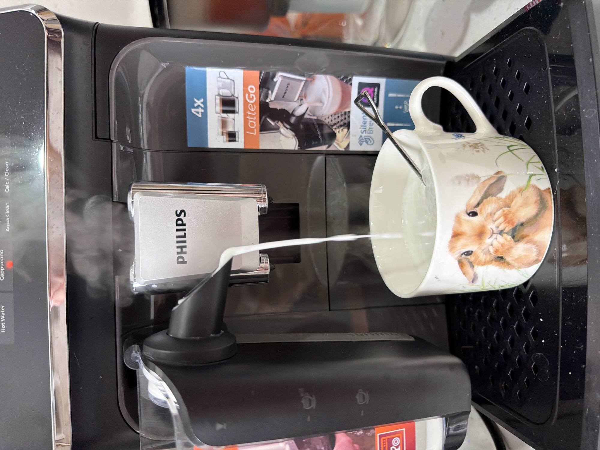 A black Philips coffee machine is dispensing milk into a white mug with a rabbit design. A spoon is also in the mug. The machine is in operation, as evidenced by the visible steam. The LatteGo branding is visible on the machine, suggesting the presence of an automatic milk frother. It is not possible to determine the city of Chaikovsky, Russia, from the visual information provided.