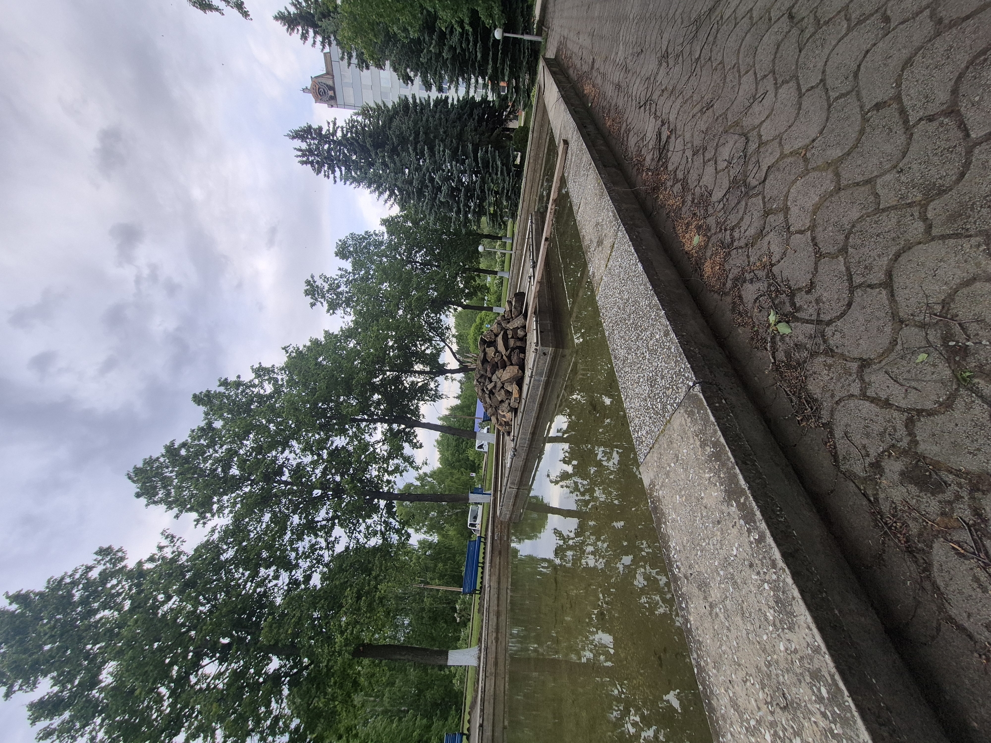 The image shows a rectangular pool in a park-like setting. The pool is bordered by concrete and contains murky water. Reflections of trees and the sky are visible in the water. A stack of rocks sits on a ledge within the pool. The edge of the pool is adjacent to a paved area with a repeating pattern. Beyond the pool, there are trees and possibly a building in the background. The image is in Chaikovsky, Russia.