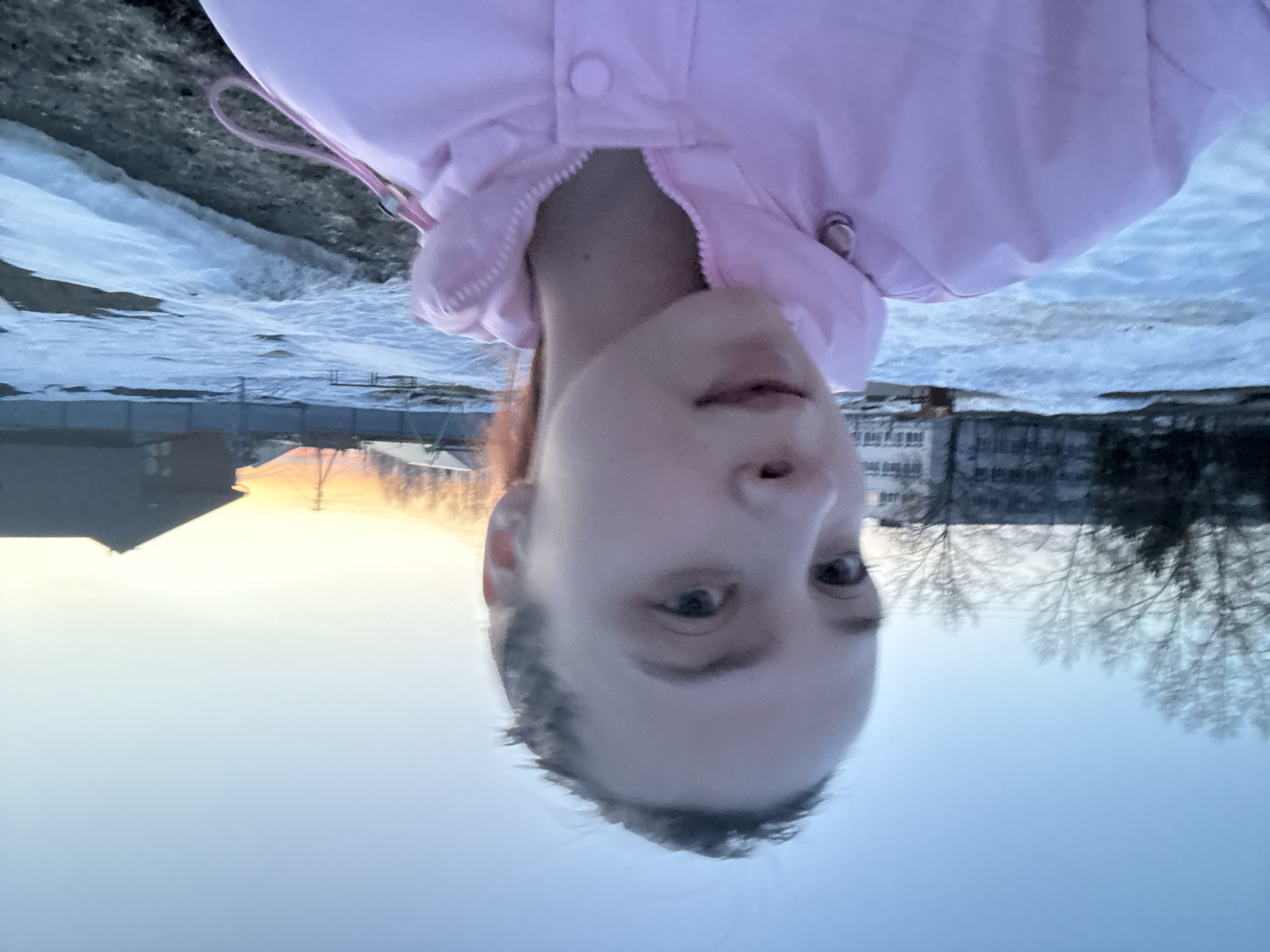 The image depicts a selfie taken outdoors in Прикамский, Russia. A person with fair skin, likely a young adult, is positioned upside down in the frame. They are wearing a pink jacket. The background shows a winter scene with patches of snow-covered ground. In the distance, there are buildings, bare trees, and the glow of the setting sun on the horizon.