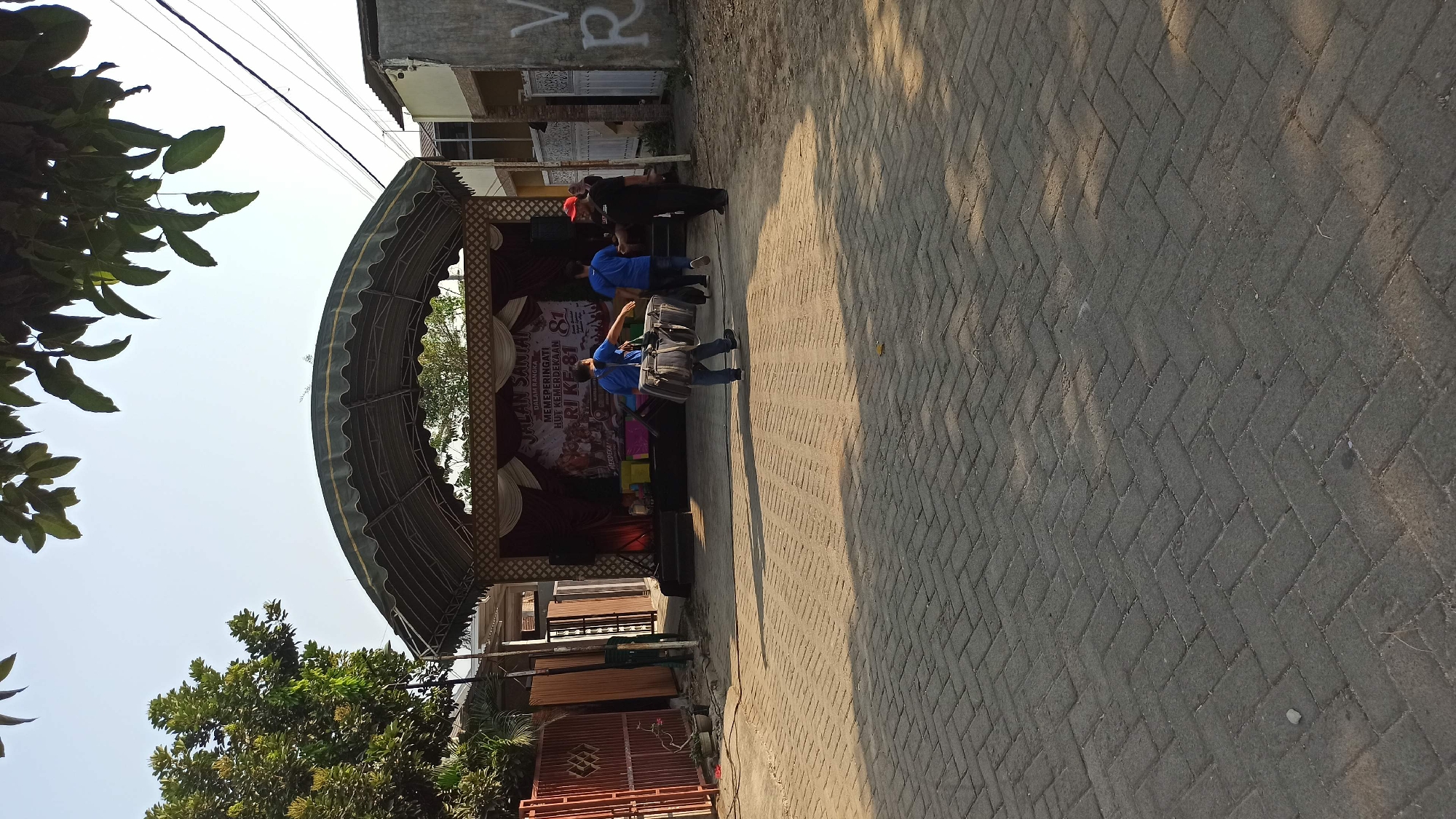 The image depicts an outdoor scene in what appears to be a street or courtyard in the city of Galak, Indonesia. The foreground is paved with grey interlocking bricks laid in a herringbone pattern, with shadows indicating the sun is at a moderate angle. In the mid-ground, there is a raised stage covered by a green arched canopy. A banner is displayed on the stage with Indonesian text, including "JALAN SANTAI MEMPERINGATI HUT KEMERDEKAAN RI KE-81," suggesting an event celebrating the 81st anniversary of Indonesian Independence.
Three individuals are visible in front of the stage. Two are wearing blue shirts and appear to be carrying equipment, possibly related to the event. One of them, facing away from the camera, is carrying a large, grey duffel bag over his shoulder. The third person, wearing a red cap and black clothing, is interacting with one of the individuals in blue.
To the left of the stage, there is a red metal fence and some potted plants. To the right, part of a building with grey concrete walls and white window frames is visible. The environment suggests a public space, potentially a community gathering or celebration.
