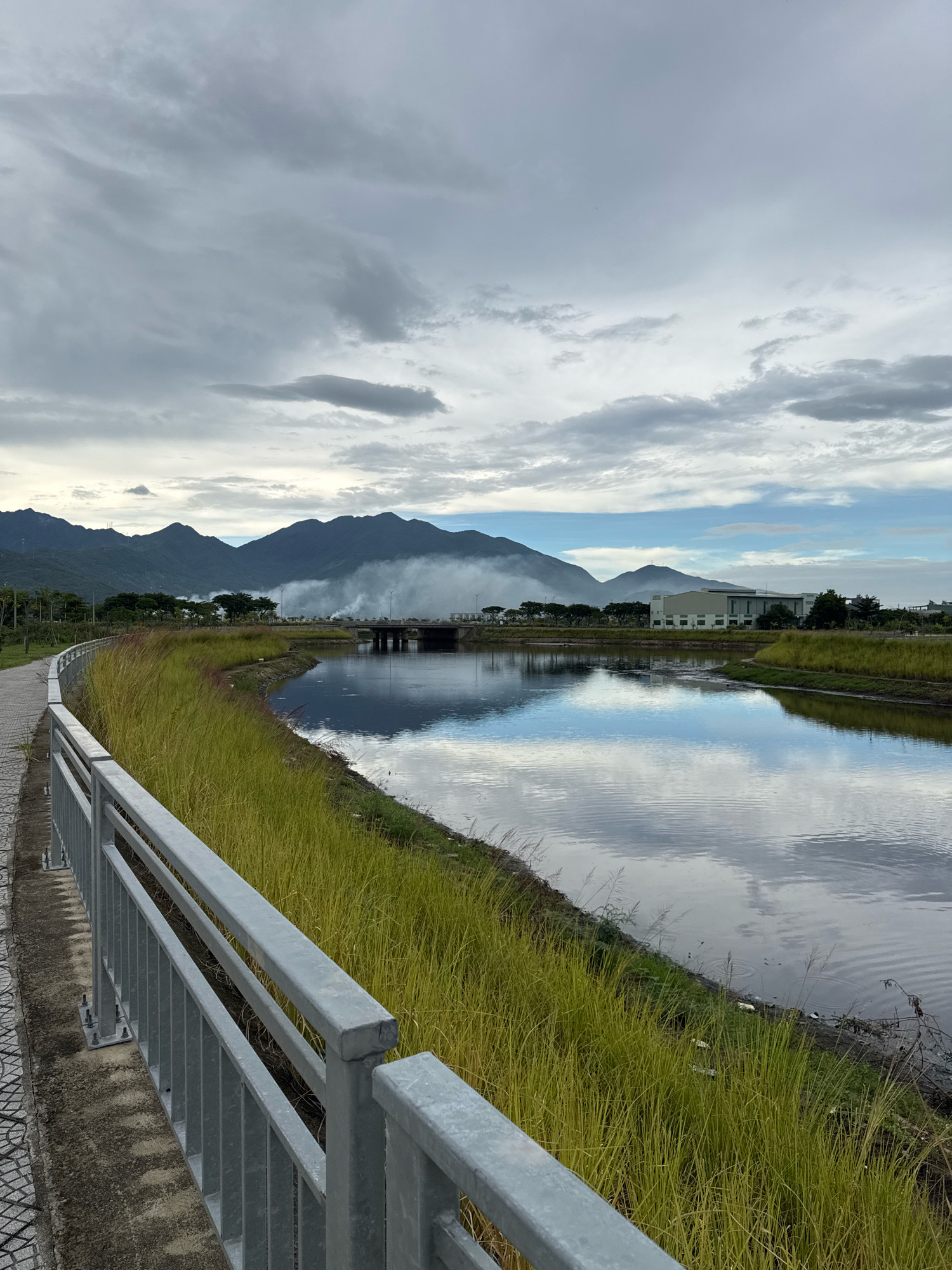 A scenic view of a body of water with mountains in the background. A paved walkway with a metal railing runs along the left side of the image, bordering tall, dry grass. The water reflects the cloudy sky and the distant mountains, creating a mirror-like effect. In the middle ground, a bridge spans the water, and a plume of white smoke rises from behind the trees on the far bank. Buildings are visible on the right side of the image, and more mountains are in the distance, partially obscured by clouds. The overall impression is one of a tranquil, natural landscape with a touch of industrial presence.