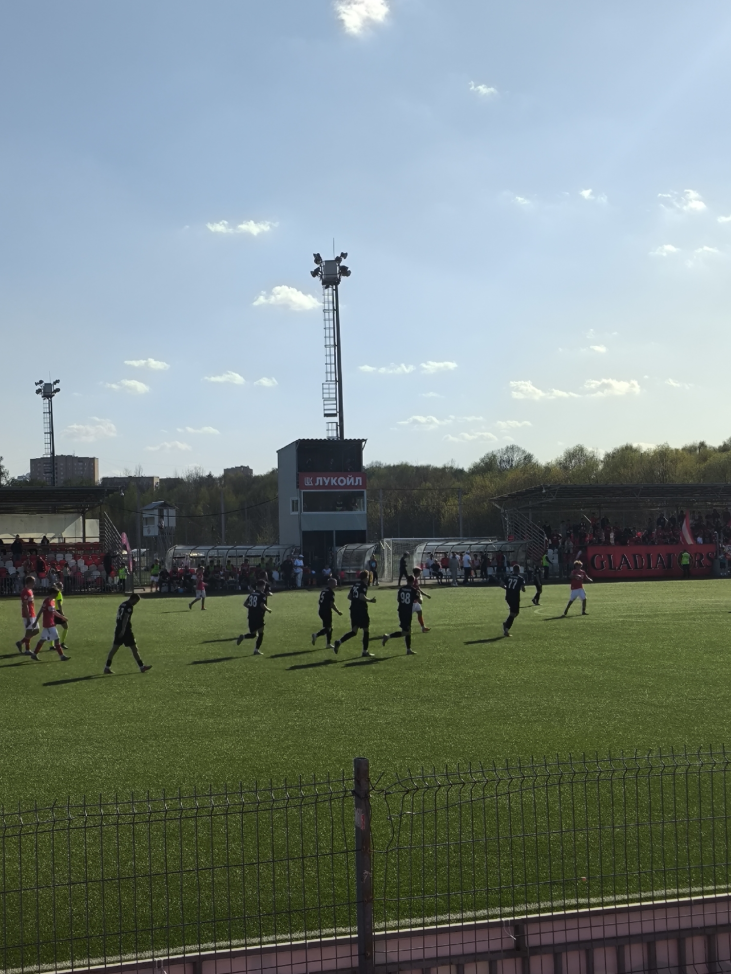 The image depicts a soccer game in progress. Players wearing red and black jerseys are actively playing on a bright green field. Spectators can be seen watching from a seating area with a banner displaying the word "GLADIATORS." In the background are stands, a tower structure with "ЛУКОЙЛ" (Lukoil) signage, trees, and buildings. The sky is blue with scattered clouds. The image appears to be taken from behind a fence. It is located in the city of Moscow, Russia.