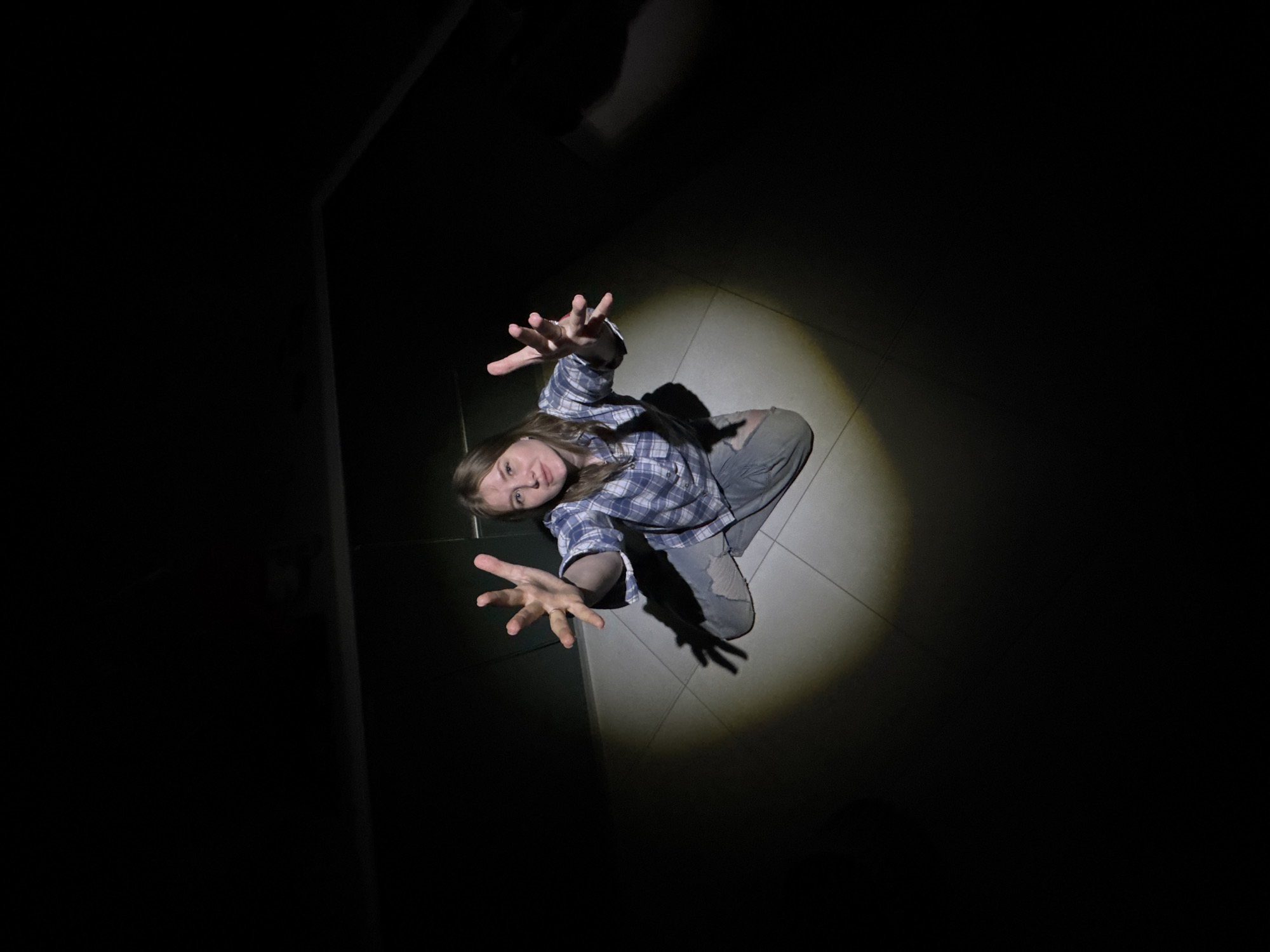 A young female kneels on a tile floor in Gornyi, Russia, arms extended. The scene is lit by a single, overhead light source. She wears a plaid shirt and ripped jeans. The surrounding area is dark and undefined. A doorway or wall edge is visible at the left of the frame.