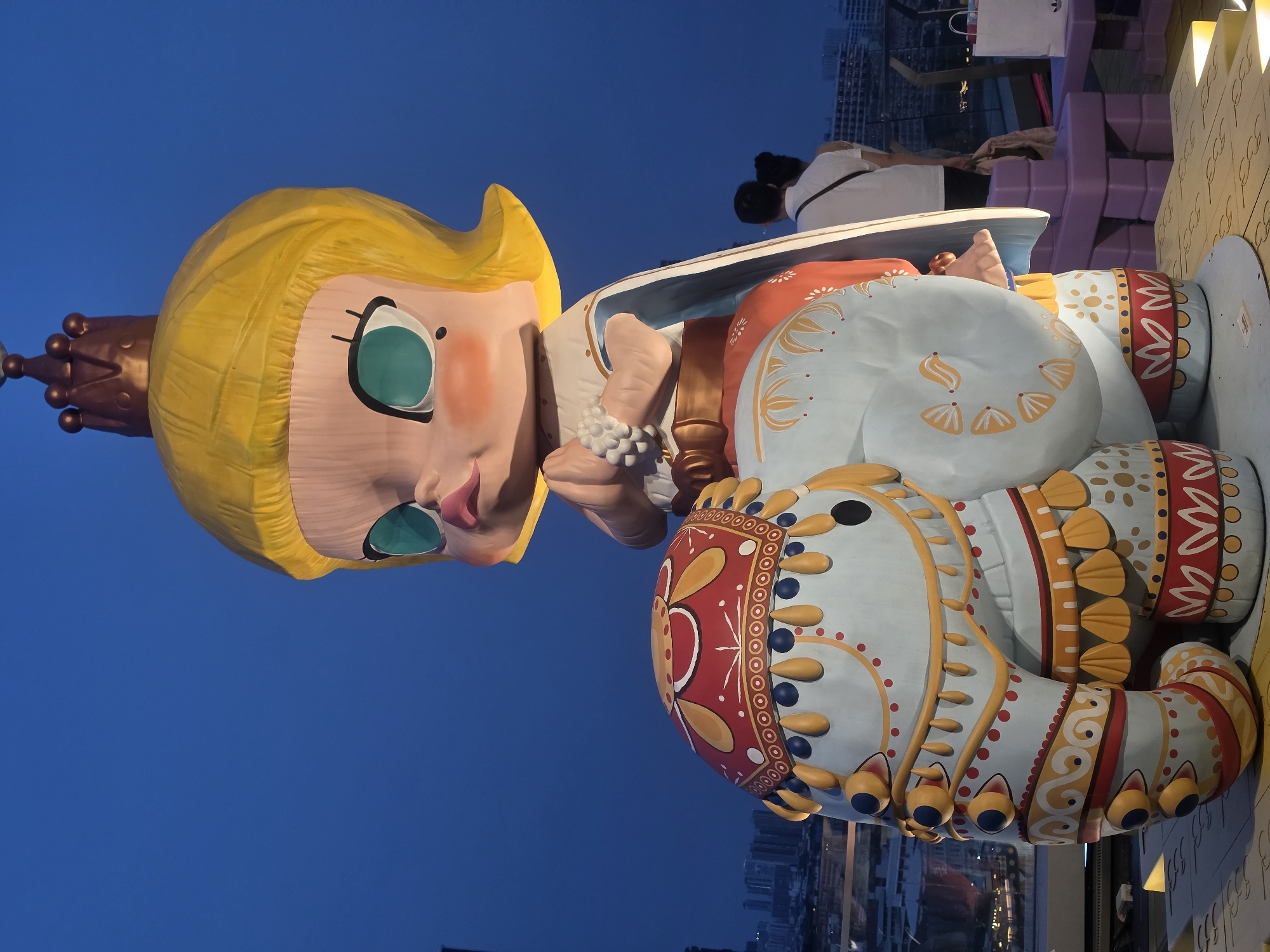 A large, colorful, seated doll-like sculpture stands outdoors, likely elevated for viewing from below. The sculpture has a cartoonish face with yellow hair, a crown, and large, light-green eyes. Its body is elaborately decorated with patterns in red, gold, and blue on a light-blue base. The background features a dusk-like, deep-blue sky and a blurred cityscape with buildings. In the upper portion of the image, behind the sculpture, there are a couple of individuals leaning over the edge of a platform. The location is likely Bangkok, Thailand, although this cannot be confirmed from the image.