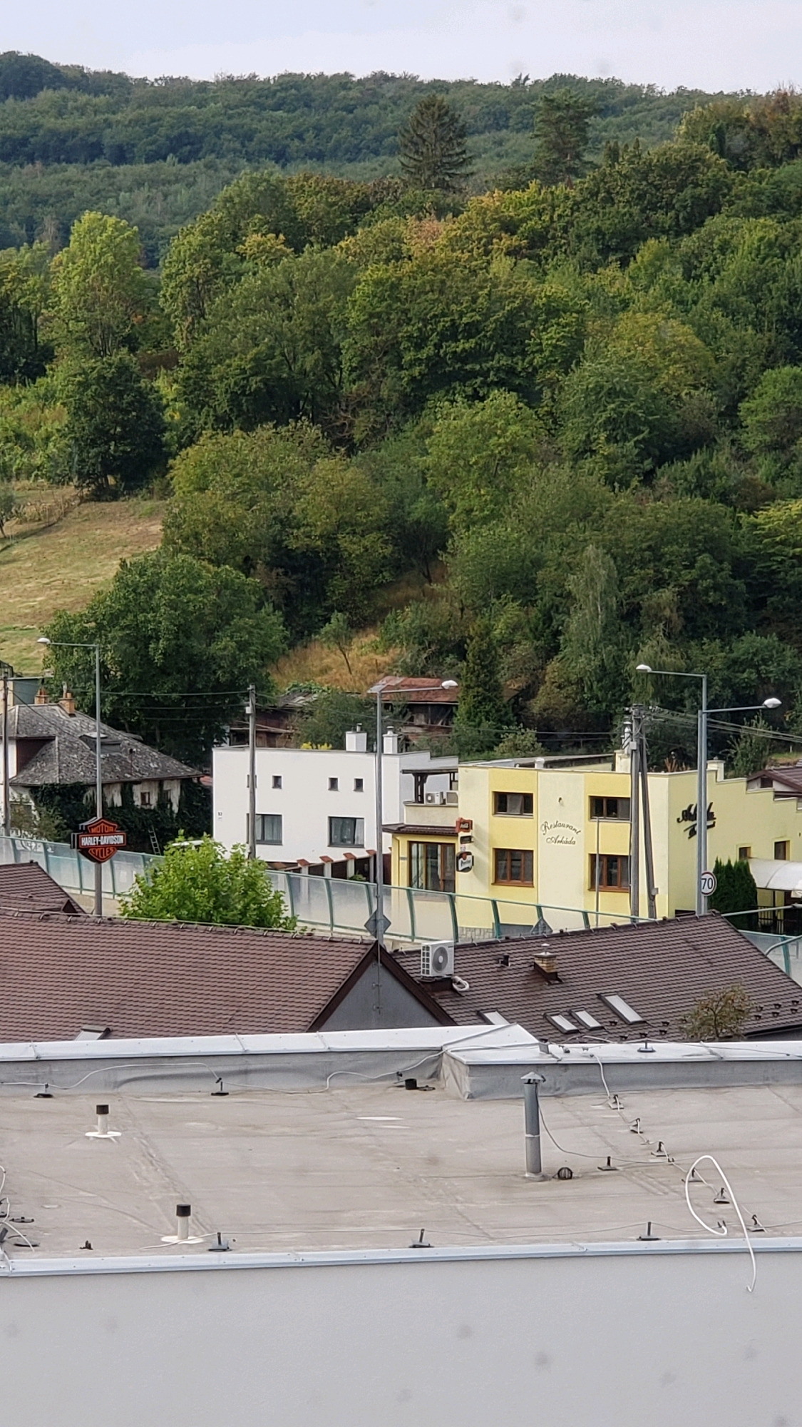 The image captures a suburban or semi-rural scene in Prešov, Slovakia. In the foreground, a flat, grey rooftop is visible, featuring several ventilation pipes and electrical conduits. Beyond this rooftop, a sloping landscape is dotted with various buildings and abundant greenery.
Prominently featured are two buildings in the mid-ground. To the left, a white, modern cubic building has several rectangular windows. To its right, a bright yellow building exhibits a more complex architectural style with multiple windows and an entrance canopy. A sign on the yellow building indicates "Restaurant Andelika". Above these buildings, a dark, sloped roof with dormer windows is visible. Further to the left, a darker, older building is partially obscured by ivy.
In the background, a densely wooded hill rises, covered in a variety of green trees. A few sparse trees are also present on a lighter, grassy slope to the left of the buildings. The sky is overcast and a pale grey. A Harley-Davidson sign is visible on a pole near the older building. The overall impression is of a residential area with commercial establishments nestled amongst the natural landscape.