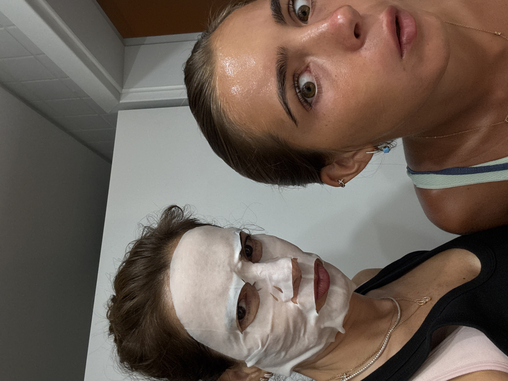 Two women are posing for a selfie in a bathroom. The woman on the left is wearing a white sheet mask that covers most of her face, revealing her eyes and lips. She has dark brown hair pulled back and is wearing a black tank top and multiple necklaces. Her expression is serious. The woman on the right, facing forward, has her eyes wide open and mouth slightly agape, with a surprised expression. She has her hair pulled back and is wearing a light blue and white striped tank top and earrings. Her face is shiny, likely from a skincare product. The background appears to be a bathroom wall with tiled sections and a doorway. The image is taken from a close-up perspective, focusing on the faces of the two women.