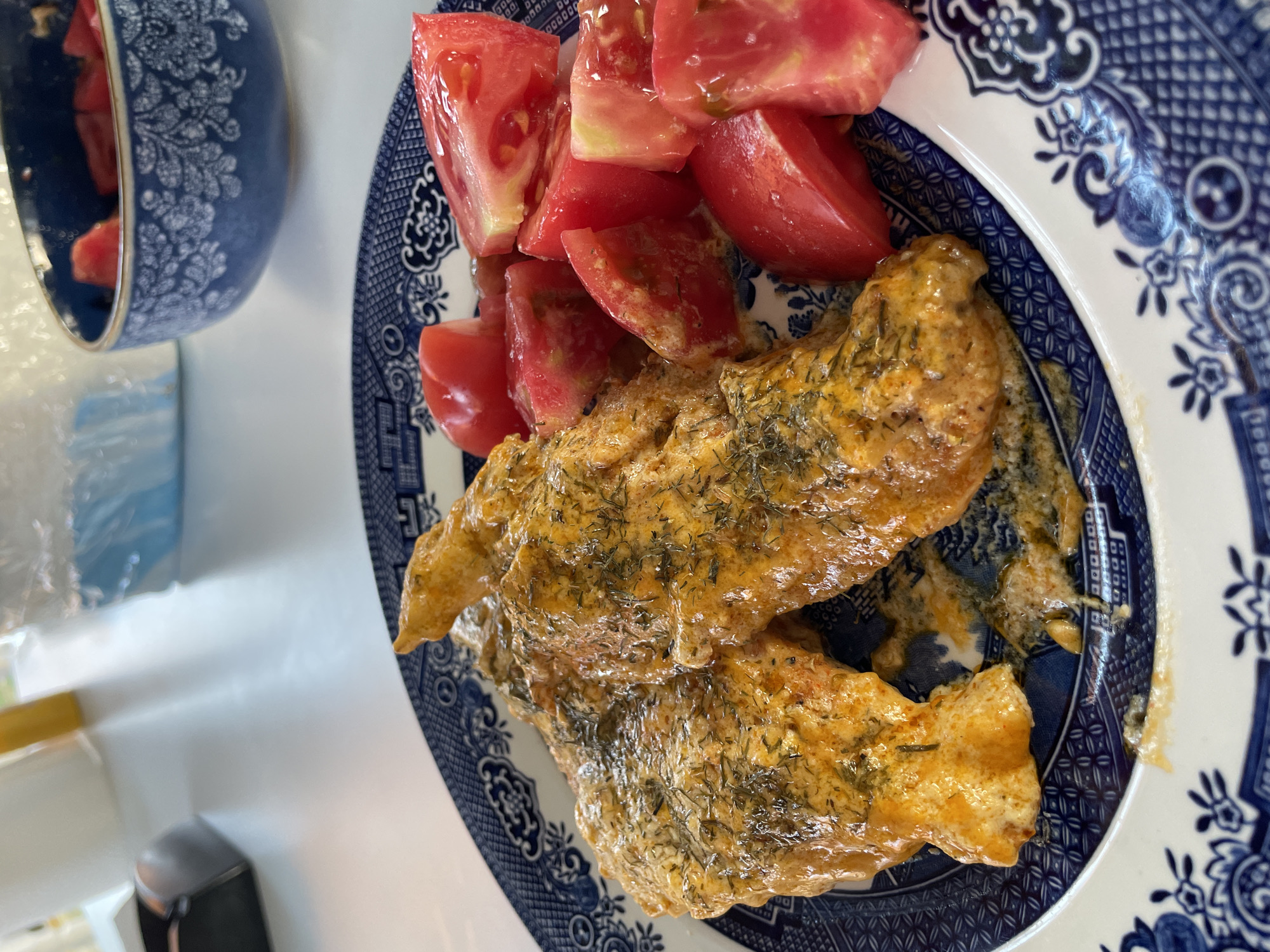 This media file features a close-up shot of a plate of food, likely a meal prepared in Skala Avdiron, Greece. The food consists of two pieces of what appears to be chicken or fish, covered in a creamy, orange-colored sauce and sprinkled with dried dill. Accompanying the main dish are several wedges of fresh, ripe tomatoes. The food is served on a blue and white patterned plate, possibly Willow pattern. In the background, a blue and white patterned bowl can be seen, also with a floral design. The setting appears to be indoors, with a white surface beneath the plate and bowl. The lighting suggests it is daytime, but there are no specific weather cues. The overall impression is of a home-cooked meal, ready to be eaten. There is no visible text in the image.