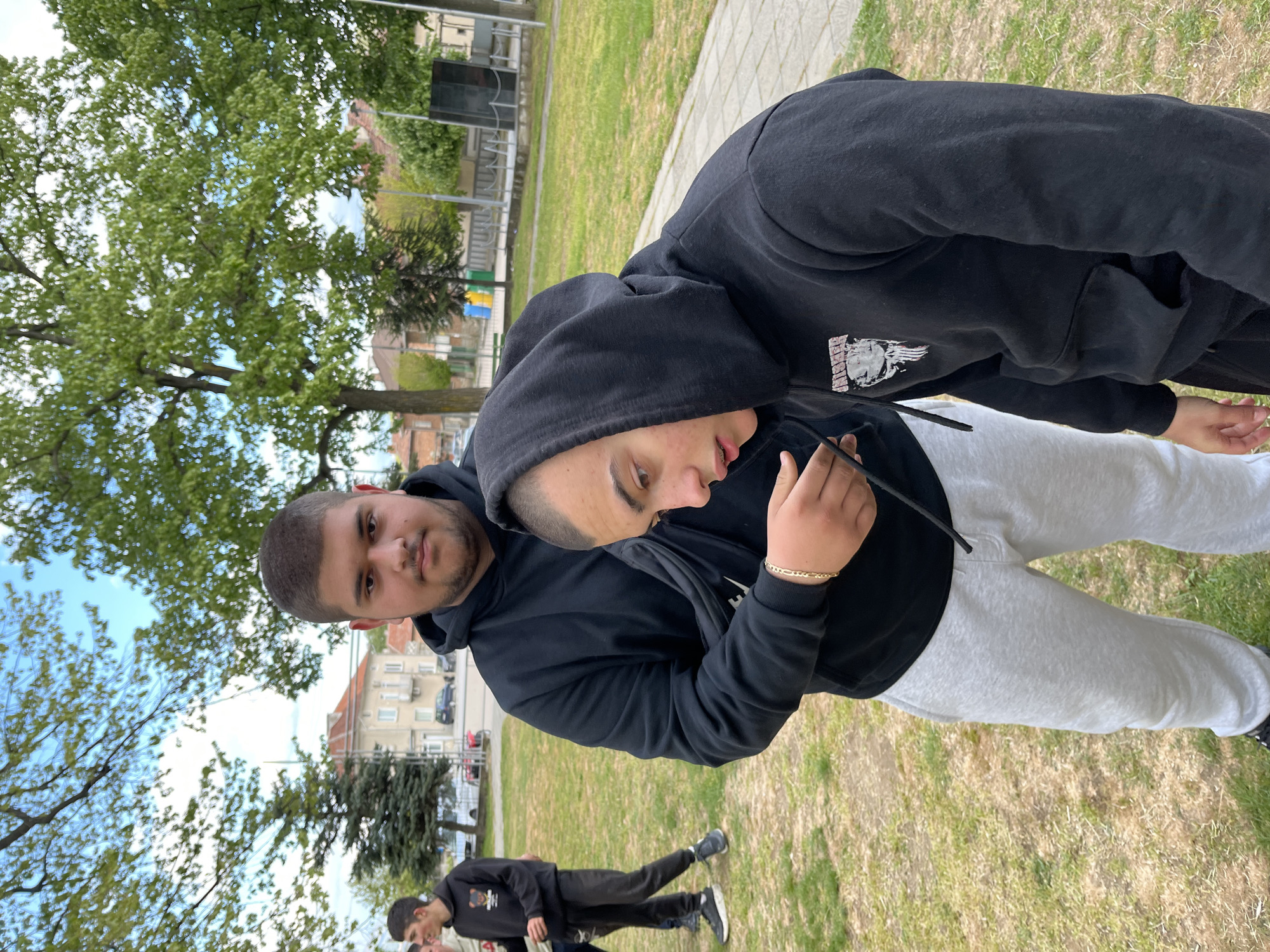 Two young men are photographed in an outdoor setting. The first man, positioned on the left, has short dark hair and a short beard. He is wearing a black hoodie. The second man is directly beside the first one. He has short dark hair, braces, and is also wearing a black hoodie with a graphic on the sleeve, along with light-gray sweatpants. The two men are standing together. In the background, there are at least two young men walking. The scene takes place in a grassy area, with trees and buildings visible in the background. The photo was taken in Karavelovo, Bulgaria.