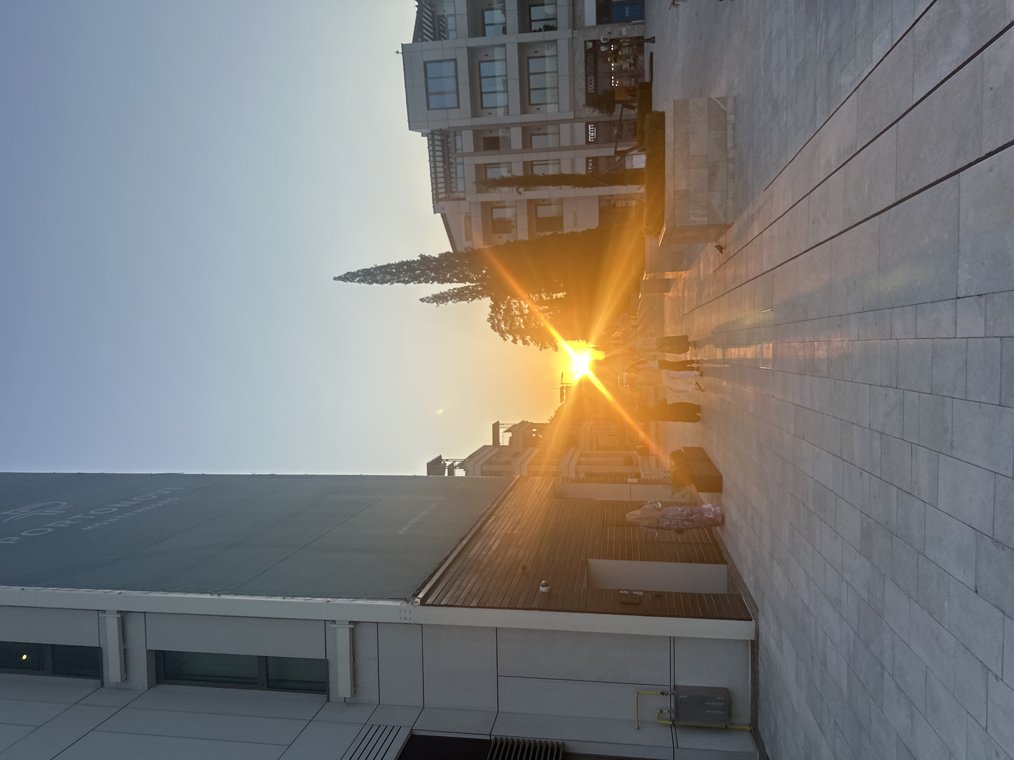 The image captures a sunset scene in the city of Kumbor, Montenegro. The sun, positioned low in the sky and slightly to the right of the frame's center, emits a strong glare, casting long shadows across the paved plaza. In the foreground, the plaza is made of large, grey square tiles. To the left, a modern building with a white facade and large windows dominates the left side of the image, with a large, grey banner on its exterior. Further into the scene, on the right, other modern buildings with multiple stories and balconies are visible. Several people, dressed in light-colored clothing, are walking on the plaza, and their figures are elongated by the backlight of the setting sun. Some of the people are wearing headscarves. A slender, tall tree stands in front of one of the buildings. The sky above is clear and transitions from a pale blue at the top to a warmer hue near the horizon, reflecting the colors of the sunset.