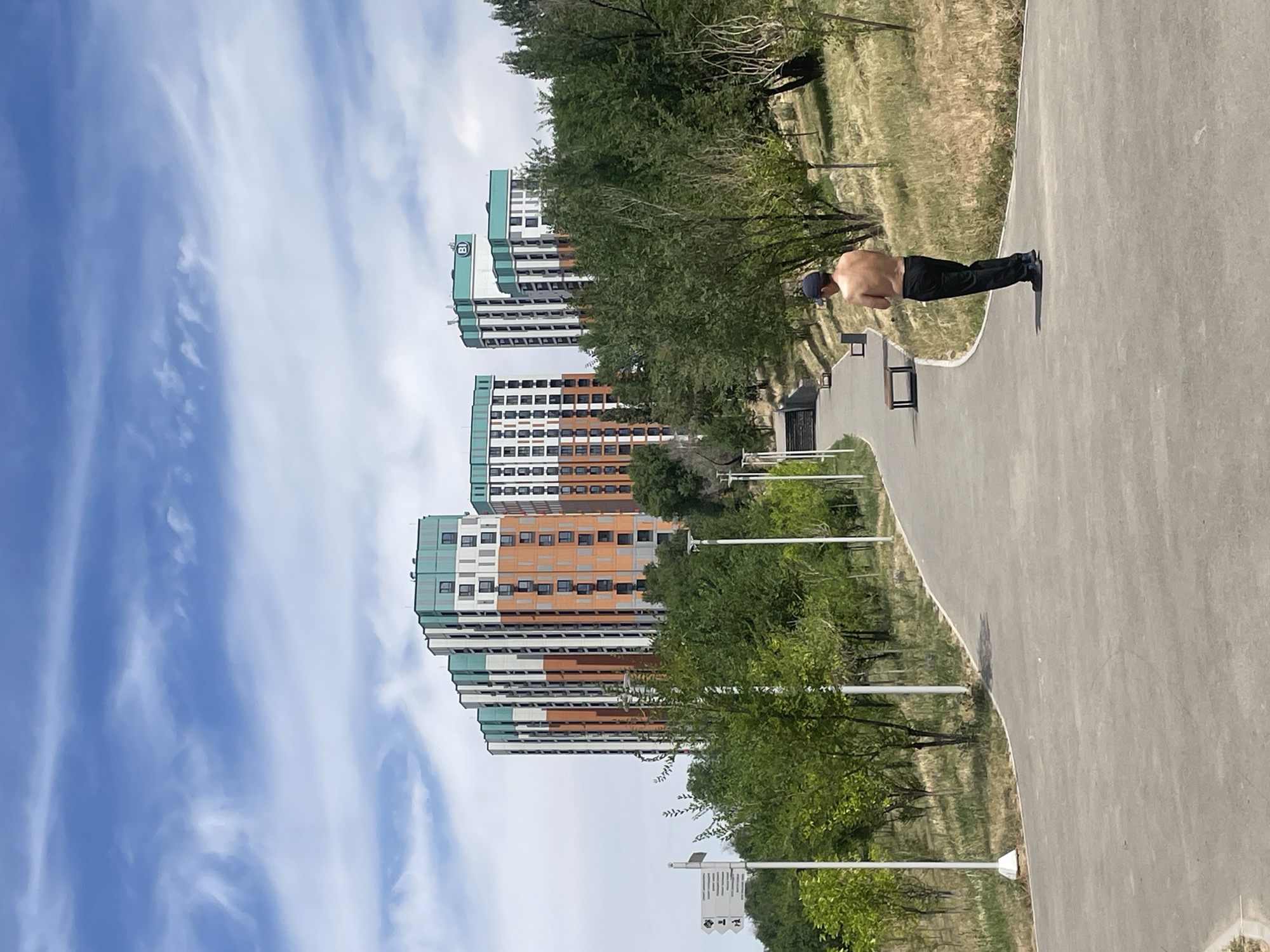 A man, shirtless and wearing dark pants and a baseball cap, stands facing away from the camera on a paved path in a park-like setting. The path curves to the left and ascends a gentle slope. To the right of the path, dry grass and scattered trees dominate the landscape. In the background, several modern, multi-story residential buildings are visible, characterized by their colorful facades featuring orange and green accents. The sky above is partly cloudy with a mix of blue sky and wispy white clouds. Park amenities, such as benches and lampposts, are also present along the path. The scene appears to be in the city of Almaty, Kazakhstan, specifically near the foot of the Alatau mountain range, a common area for residential development and recreational spaces.