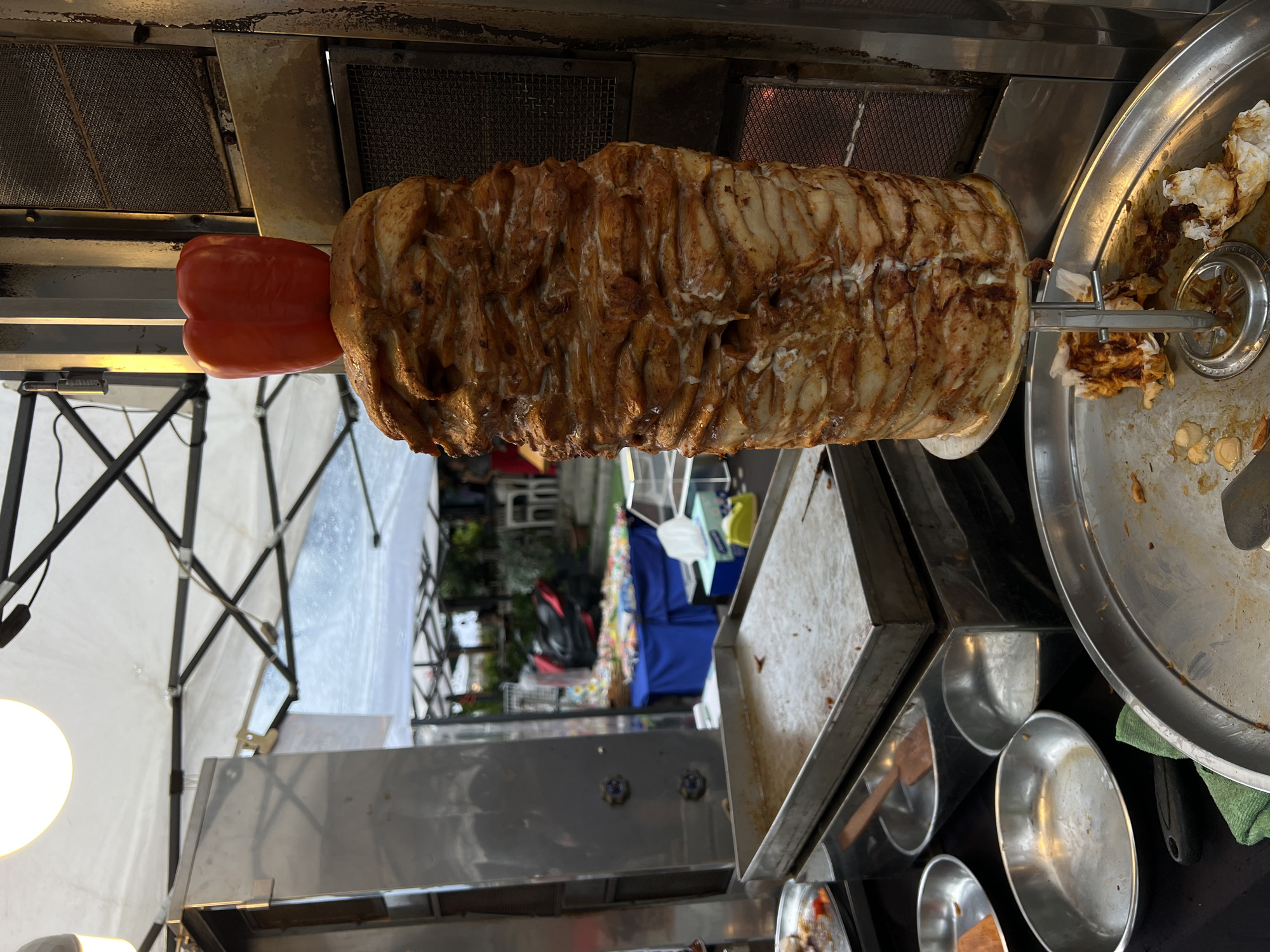The image shows a close-up view of a vertical rotisserie with a large stack of thinly sliced meat, likely chicken or lamb, being cooked. A bright red bell pepper is skewered onto the same spit, positioned to the left of the meat. The meat has a golden-brown, slightly charred appearance from the heat of the rotisserie. The scene appears to be outdoors, possibly at a food stall or market, indicated by the presence of a white tent structure with metal poles in the background. There are also some stainless steel surfaces visible, suggesting a food preparation area. The overall impression is one of a food preparation setting, with the focus on the cooking meat. The lighting suggests it might be daytime, but the specific time is not clear. There is no visible text in the image. The crops highlight different sections of the rotisserie and surrounding equipment.