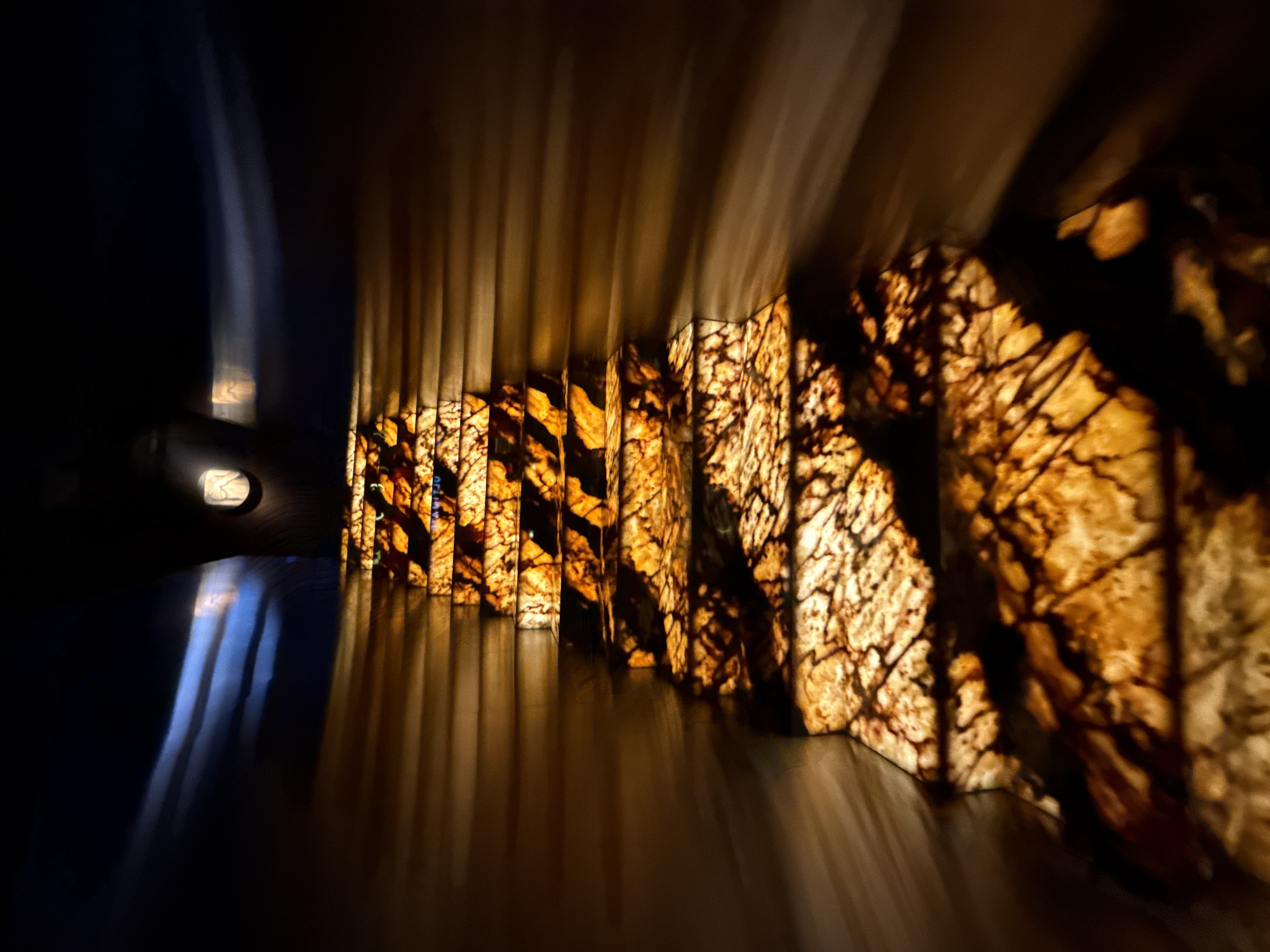 This abstract interior shot, set in Moscow, Russia, captures a dramatic interplay of light and shadow within what appears to be a decorative architectural space or a well-lit hallway. The primary subjects are a series of vertically arranged, illuminated panels on the right side of the frame. These panels are translucent, glowing with a warm, golden-brown light that reveals intricate, dark, organic patterns, reminiscent of natural veins in stone like onyx or marble, or possibly stylized tree branches. The light casts long, vertical reflections and streaks onto a dark, reflective floor.
The left side of the image is largely enveloped in deep shadow, providing a stark contrast to the illuminated panels. Within this dark expanse, soft, vertical streaks of bluish-white light are visible, along with a small, circular light source or reflection positioned higher up on the left. The perspective and streaky lighting suggest a sense of depth and movement, or perhaps a long exposure effect, emphasizing the linear elements.
No people or specific activities are depicted in the scene. The artificial interior lighting makes the time of day or weather indeterminate. The overall atmosphere is one of modern elegance, with a focus on texture, light, and abstract forms, evoking a sense of calm and visual intrigue. There is no visible text.