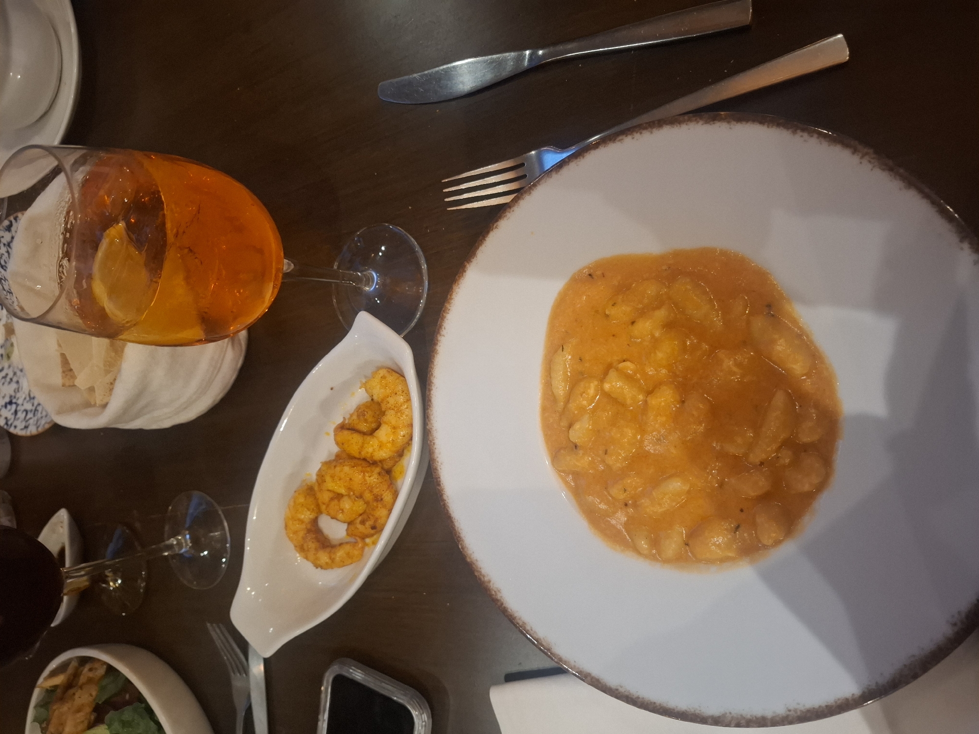 The image shows a restaurant table setting with a plate of gnocchi in an orange sauce, a small dish of spiced shrimp, a glass of Aperol spritz with a lemon slice, bread, a salad, a fork, and a knife. A mobile phone is also on the table. The location is inferred to be in Jesús del Monte, Mexico.