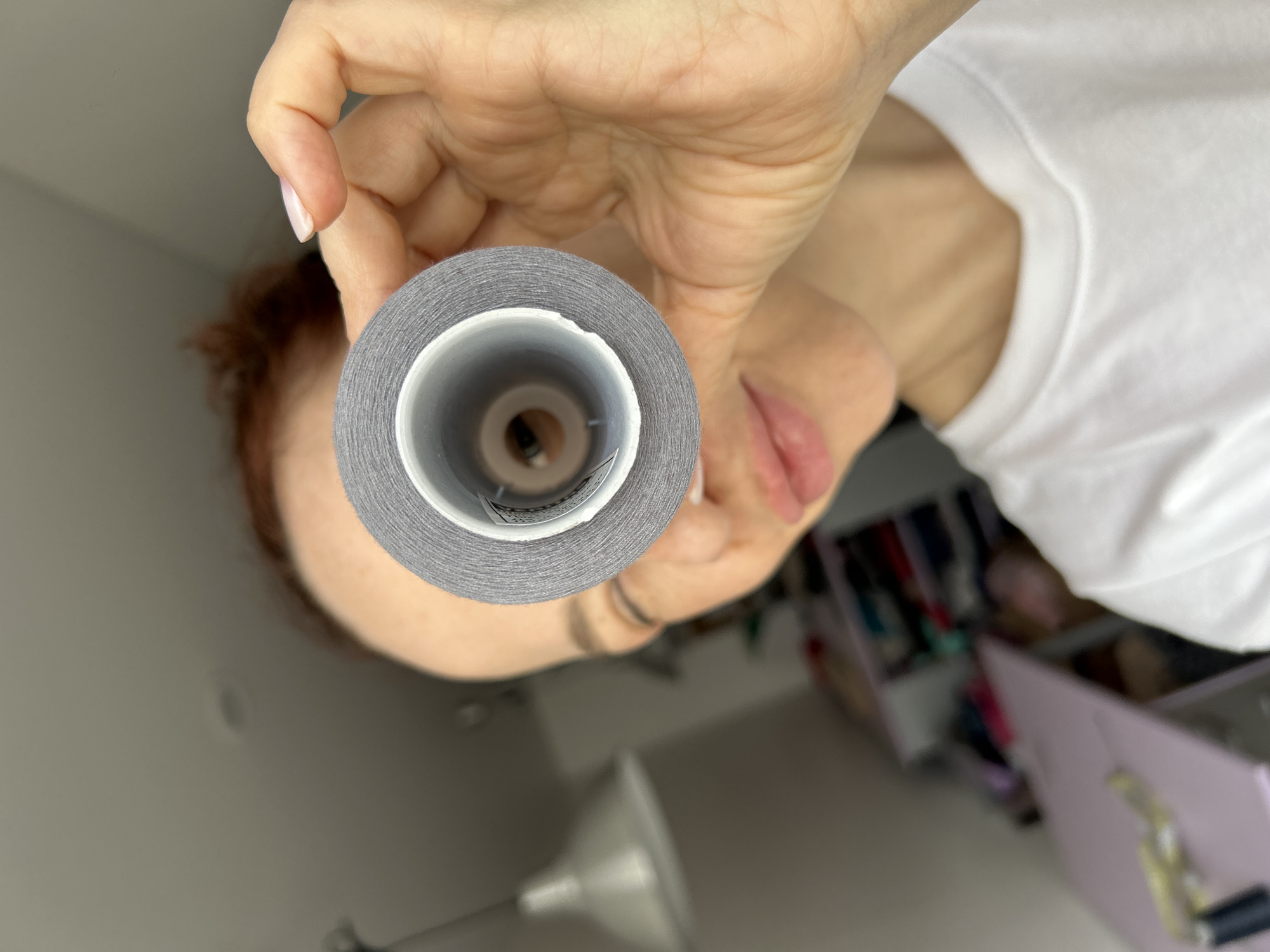 A woman with light skin and reddish hair is holding a spool of gray thread directly in front of the camera, obscuring most of her face. She has on a white t-shirt. In the blurry background, there's a glimpse of what appears to be a cluttered room with drawers, possibly in a home. The exact location is unknown, and while Chaikovsky, Russia, may be relevant context, there's no information in the image to confirm that.