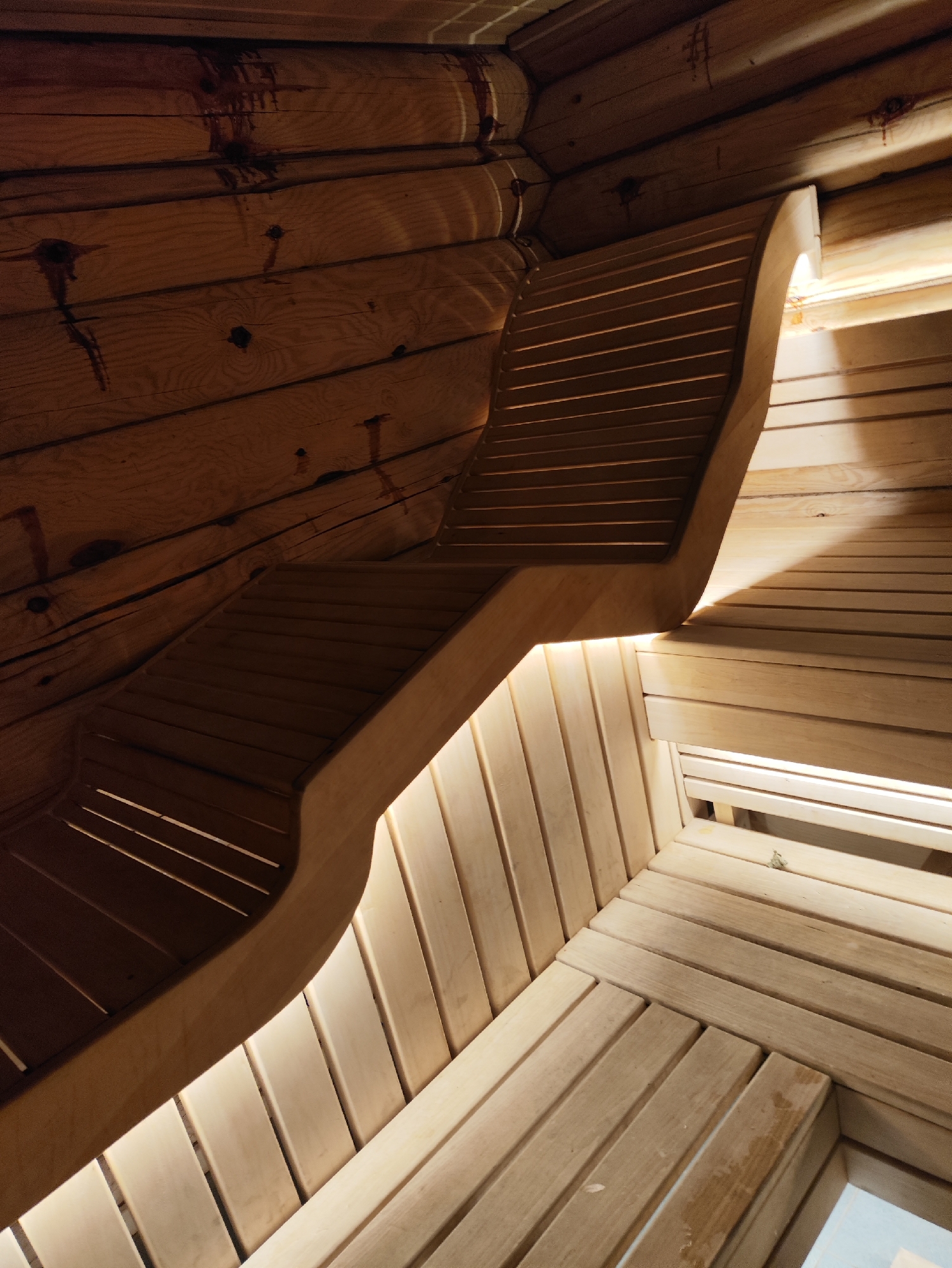 An interior view of a sauna in Старое Мартьяново, Russia, showcases the warm tones of wooden log walls and slatted wooden benches. The benches are illuminated by soft, warm lighting, creating a cozy and inviting atmosphere. Some of the log walls show dark stains, possibly from sap or natural wood variations. The perspective is from a low angle, looking up towards the ceiling and along the curved benches.