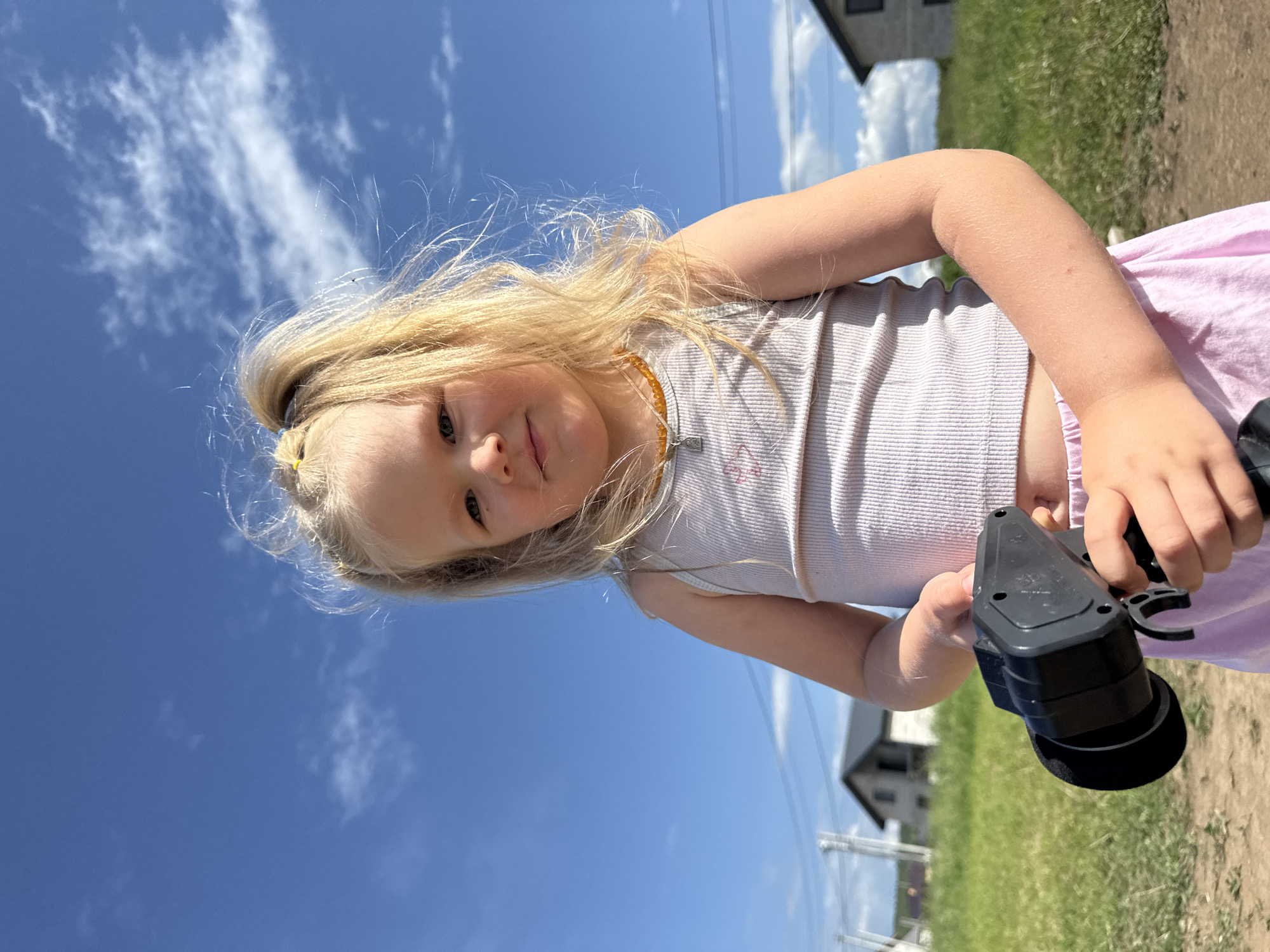 A young, blonde-haired child is standing outdoors in Ферма, Russia. She is wearing a white tank top, pink shorts, and an amber-colored necklace. She is holding a black, plastic object in her hands. The background includes a blue sky with clouds, green grass, a dirt path, and a house with a gray roof. There are also power lines visible in the distance. The scene appears to be a sunny day.