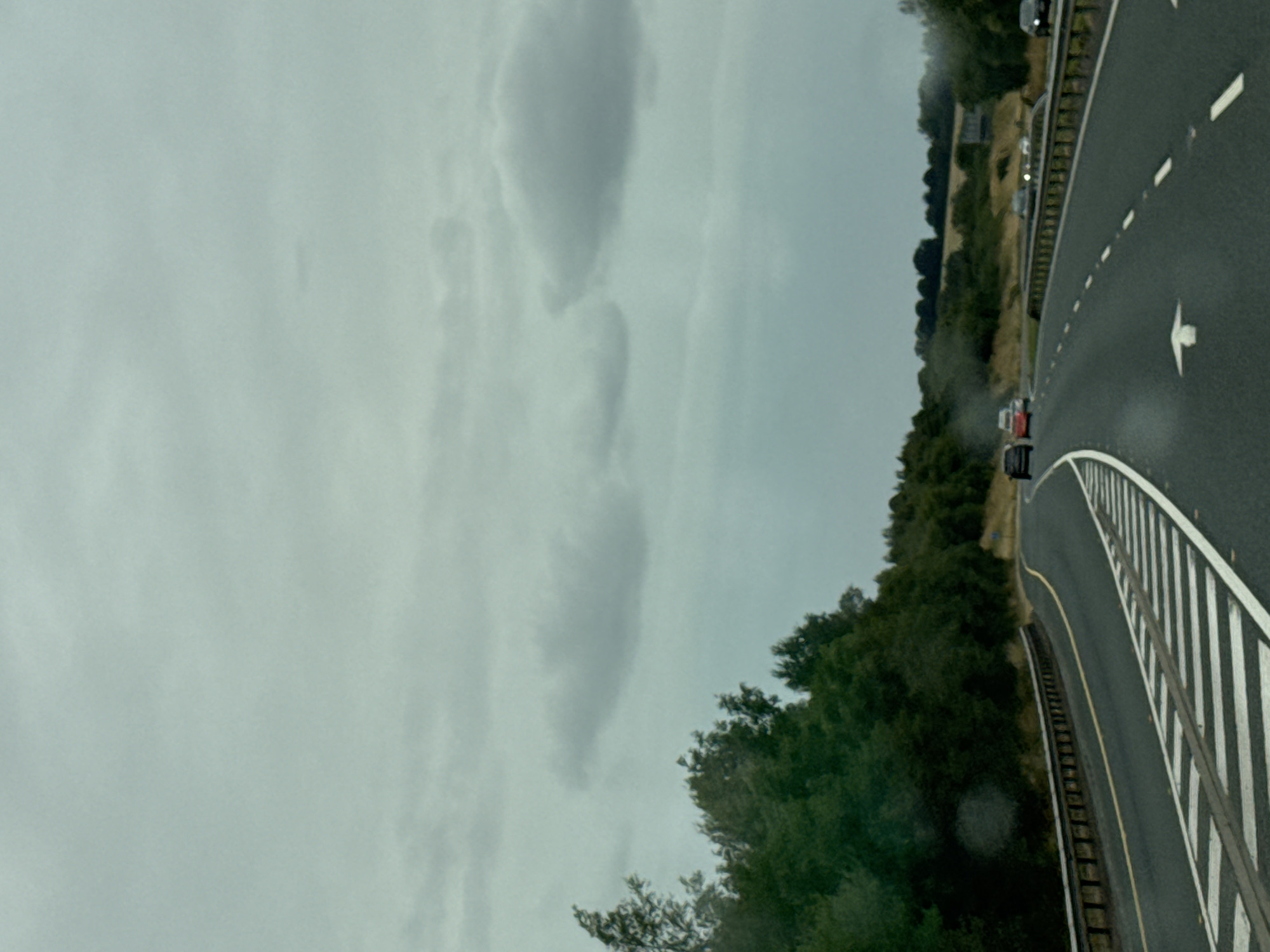 This image captures a view from a car traveling on a highway in Knockraheen, Ireland. The road is asphalt with white lane markings, including a directional arrow pointing forward. Several cars are visible on the road, some in the foreground and others in the distance. On the left side of the road, a metal guardrail runs alongside a dense line of dark green trees. The background features a gentle slope of land with scattered trees and a building. The sky is overcast with gray clouds, suggesting a cloudy day.