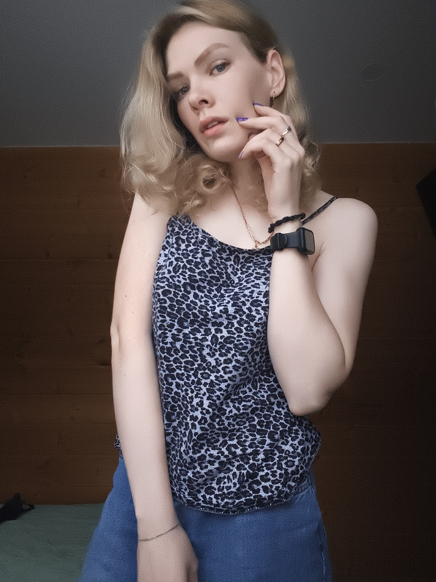 A fair-skinned woman with blonde, shoulder-length wavy hair poses indoors. She wears a blue and black animal print tank top and blue denim shorts. A black smartwatch and a beaded bracelet adorn her left wrist, and she wears rings on the fingers of her right hand. Her fingernails are painted purple. The background includes a light brown wooden paneling and a dark ceiling, with the edge of a bed visible at the bottom left. The location appears to be an indoor setting, possibly a bedroom. There is no evidence to indicate the location is the city of Русский Вожой, Russia.