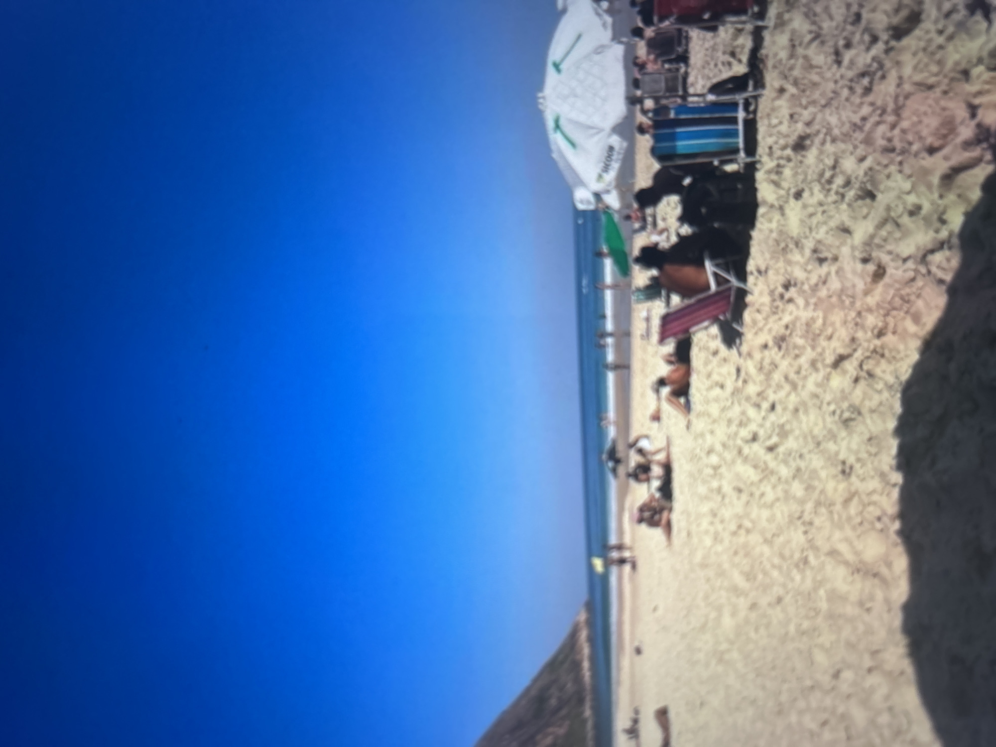 A wide shot captures a crowded beach under a clear blue sky. The sand is light brown, with people scattered across it, some lying down and others sitting in beach chairs. There are several beach umbrellas providing shade, one large white umbrella with green markings, and a smaller green umbrella. In the distance, the ocean meets the horizon, with small waves visible. To the left, a large, sloping mountain rises from the coastline. The scene appears to be in a tropical or subtropical location.