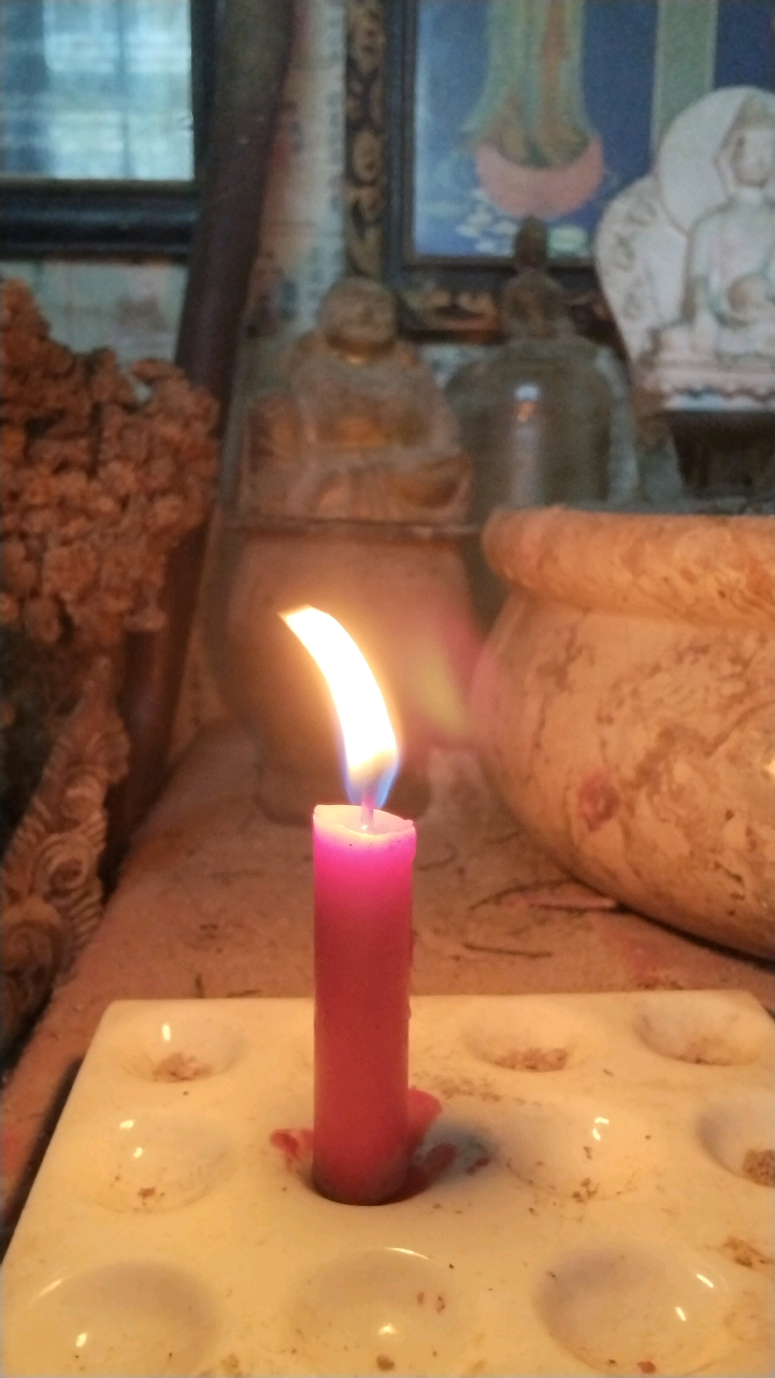 A lit pink candle stands in a white tray with indentations. The candle is centrally placed and its flame casts a warm glow on its surroundings. The background is blurred but suggests a shrine or altar with statues and artwork. The overall scene appears to be indoors. There is no discernible information regarding the city of Penompo, Indonesia, or any people.