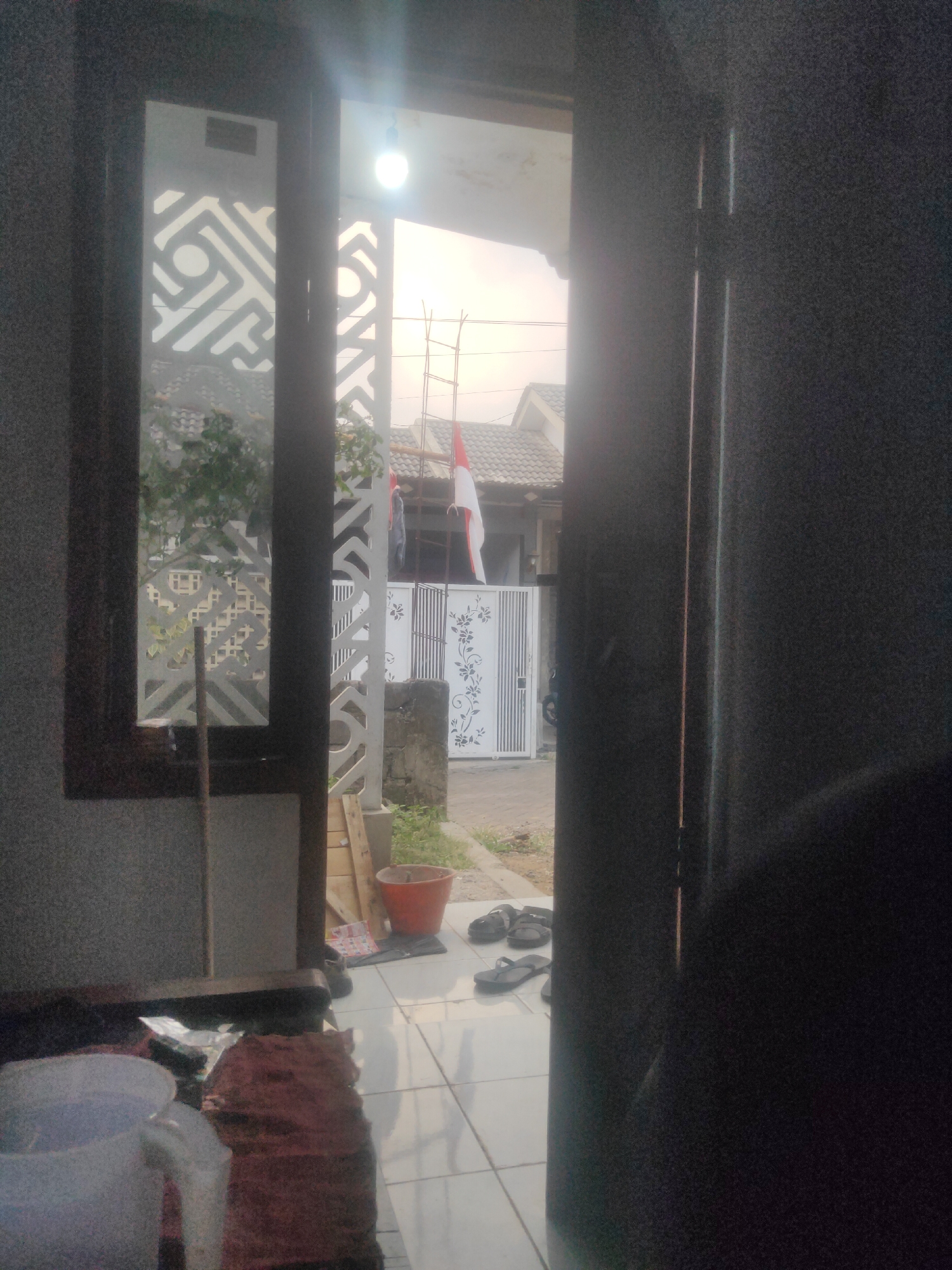 The image shows a view from inside a building, looking out through an open doorway and a window. The scene appears to be a residential area, possibly in Antirogo, Indonesia, given the presence of what looks like an Indonesian flag.
In the foreground, inside the building, there are some household items on the floor, including a white plastic bucket, a red bucket, and some dark-colored footwear (flip-flops). The floor is tiled.
Looking through the window and doorway, we see an outdoor scene. There is a white gate with a floral pattern and what appears to be a construction scaffold. An Indonesian flag, red and white, is prominently displayed. Beyond the gate, there are houses with tiled roofs. Trees and greenery are visible. The sky suggests it might be daytime, possibly late afternoon or early morning due to the lighting.
A person is partially visible in the doorway, appearing to be walking or standing near the edge of the frame, with their back to the camera. The overall impression is that of a domestic setting in a neighborhood.