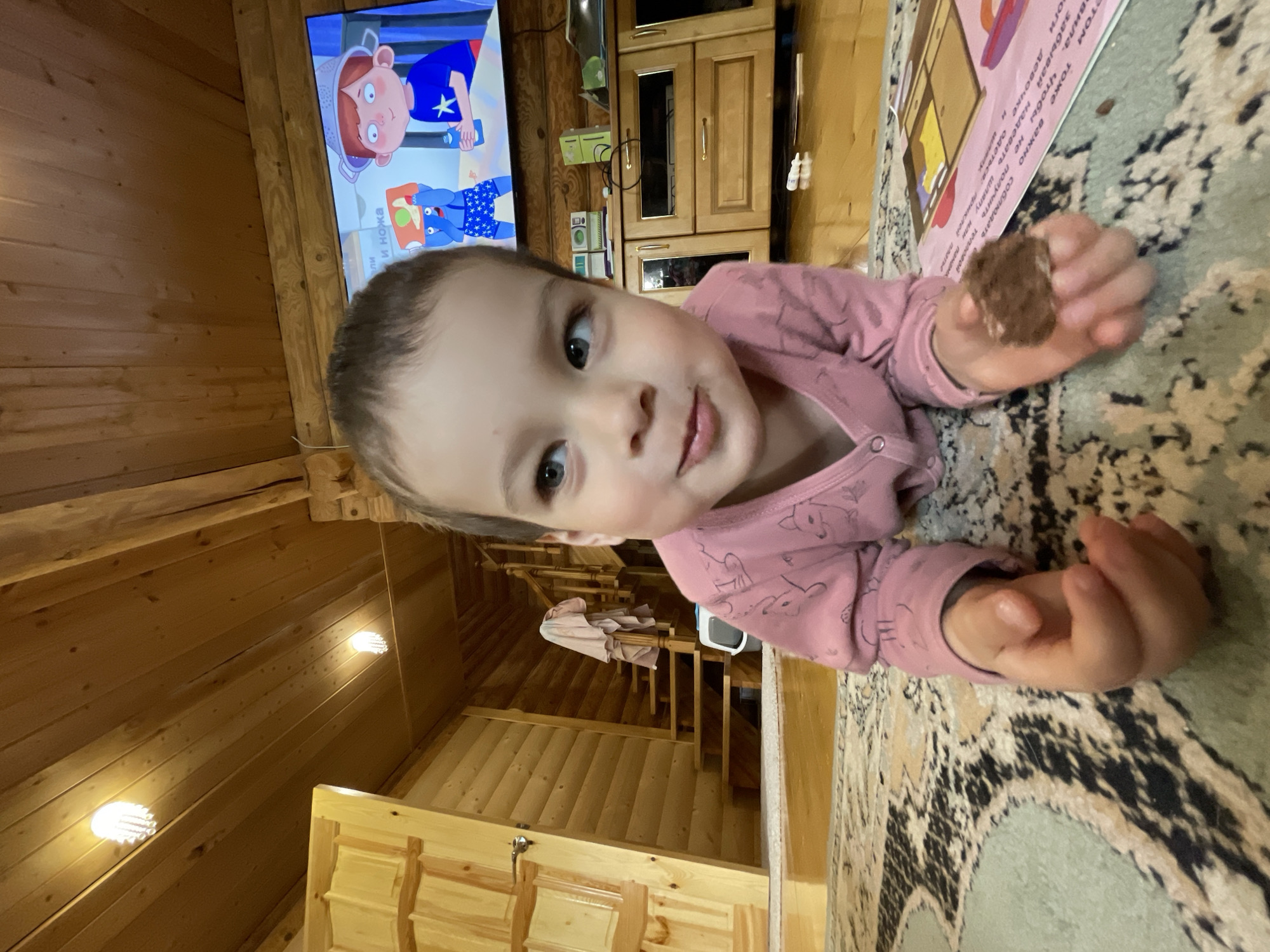 A young child wearing a pink, long-sleeved shirt with animal prints is lying on a patterned carpet, holding a piece of chocolate in their right hand and looking directly at the camera. The child is indoors, presumably in a wooden cabin, given the wooden walls and ceiling. In the background, a television shows a cartoon, and there's a wooden cabinet near the television. Stairs leading to another level of the cabin are also visible. The carpet has a textured, organic pattern of colors including beige, brown, and green. Text, potentially from a book, is visible along the upper edge of the carpet. The setting may be in Zav'yalovo, Russia, given additional information.