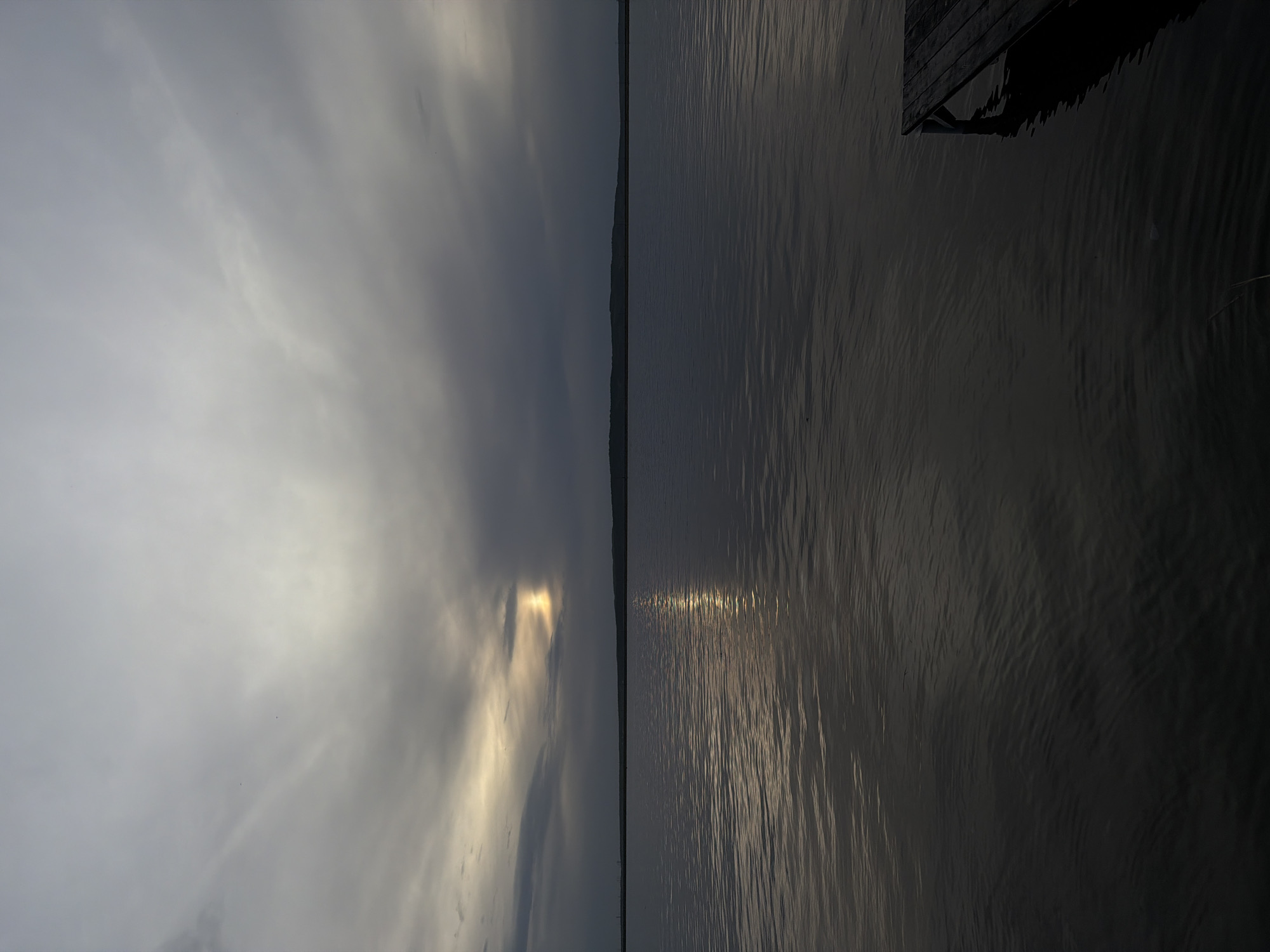 The image depicts a body of water, likely the Kama River, near the city of Chaikovsky, Russia. The left portion of the frame features a cloudy sky, with a patch of sunlight breaking through. This sunlight reflects off the water's surface, creating a glimmering effect. To the right, a dark, shadowed area appears to be a wooden structure extending over the water, possibly a pier or dock. The water's surface shows subtle ripples, suggesting a slight breeze. The overall scene is serene and natural, with a contrast between the bright sky and the dark water.