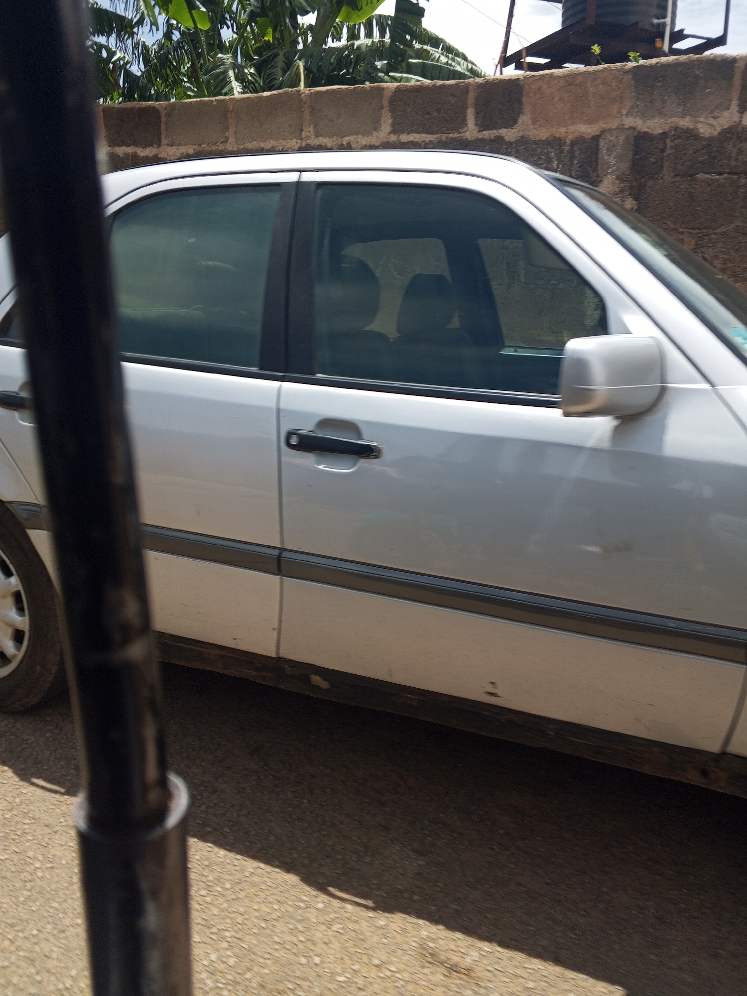 The image depicts a static outdoor scene in Jos, Nigeria, primarily featuring the driver's side of a silver Mercedes-Benz C-Class sedan, consistent with a W202 generation model. A dark, vertical pole obscures the left portion of the car, including its front quarter panel and a silver multi-spoke wheel. The vehicle displays four doors with black recessed handles and dark-tinted windows, through which a dark interior is vaguely discernible. A dark protective trim strip extends along the lower body line, and the lower panels of the car are visibly soiled with dirt and discoloration.
Behind the vehicle, a low wall constructed from reddish-brown concrete blocks is present, unevenly laid. Above this wall, dense green tropical foliage, consistent with banana plants, is visible against a light blue sky with scattered clouds. In the upper right corner of the frame, an elevated dark water tank, supported by a metal frame, is situated, along with faint utility lines. The ground beneath the car and extending into the lower right is a light brown, textured surface, appearing to be an unpaved road or dusty ground, with distinct shadows cast by direct sunlight. No individuals or active interactions are visible within the scene.