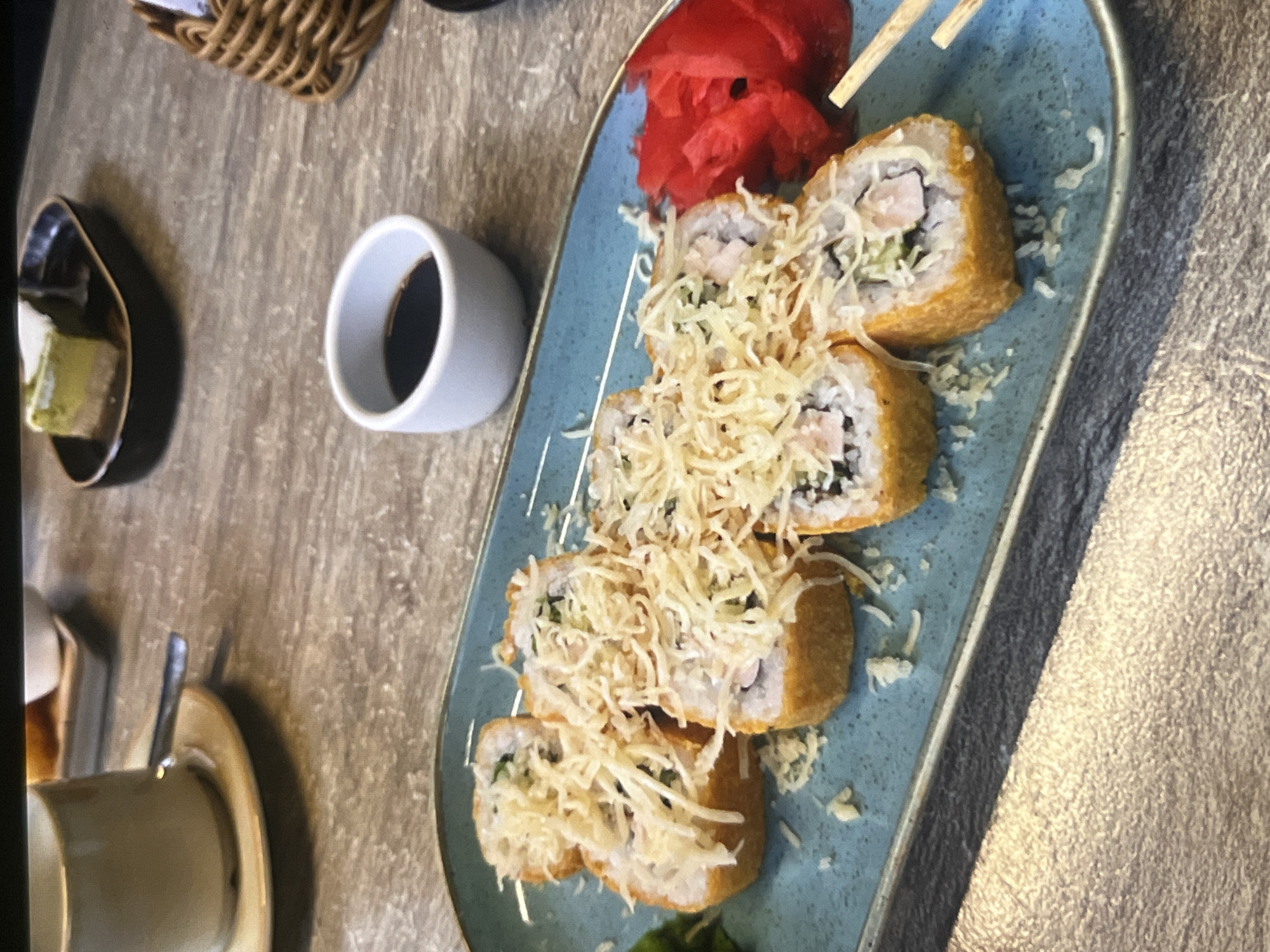 A close-up, eye-level shot captures a plate of sushi rolls on a blue rectangular platter. The sushi appears to be deep-fried, with a crispy, golden-brown exterior and topped with shredded cheese and possibly tempura flakes. The rolls are arranged in two rows on the platter. To the right of the sushi, a pile of bright pink pickled ginger is visible, along with a pair of wooden chopsticks. In the background, a small white dish of soy sauce sits on a wooden table, alongside a cup and saucer containing a beverage. A slice of green cake is also present in the background on a dark plate. The setting appears to be a restaurant.
