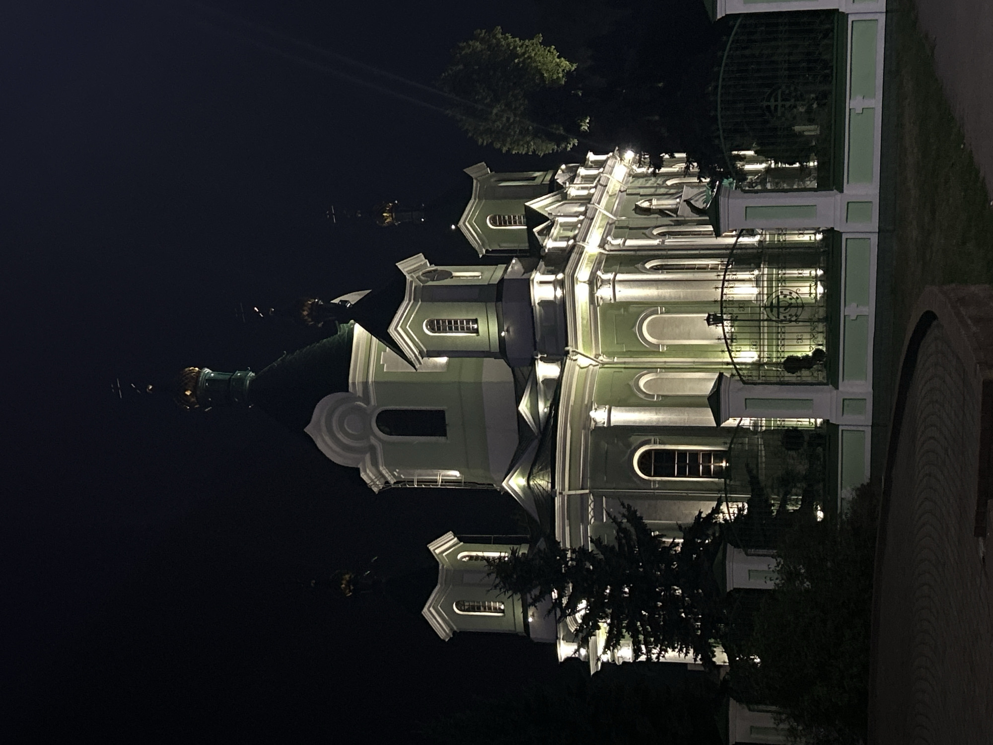 The image depicts an illuminated orthodox church at night in the city of Brest, Belarus. The church is a large, multi-domed structure with a pale green facade and white architectural details. It is lit from below by numerous spotlights, creating a dramatic effect against the dark night sky. Several trees, including a prominent evergreen tree on the left, are visible in the foreground and around the church. A decorative metal fence with ornate pillars surrounds the church grounds. The ground in the foreground appears to be paved with bricks in a circular pattern. No people are visible in the scene.