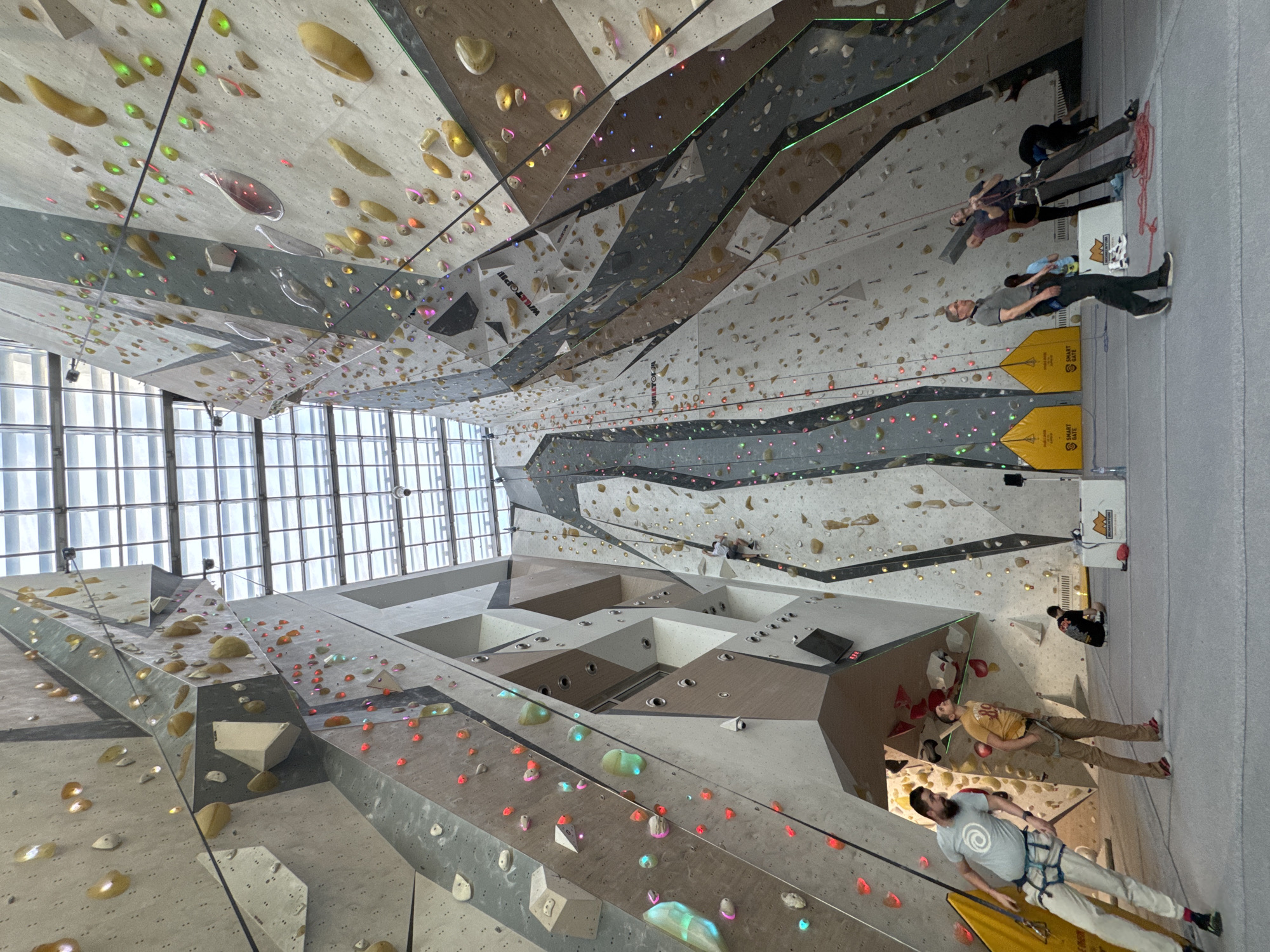 The image depicts the interior of a climbing gym with tall artificial climbing walls. Multiple individuals are engaged in rock climbing activities, some wearing harnesses and ropes. The walls are textured and feature numerous climbing holds of various shapes and colors. The facility has a modern design with visible metal beams and large windows providing natural light. The location appears to be in an urban area, given the presence of buildings visible through the windows. While the exact address is not evident from the image, the city is identified as Sofia, Bulgaria. The overall setting is a recreational indoor sports facility.