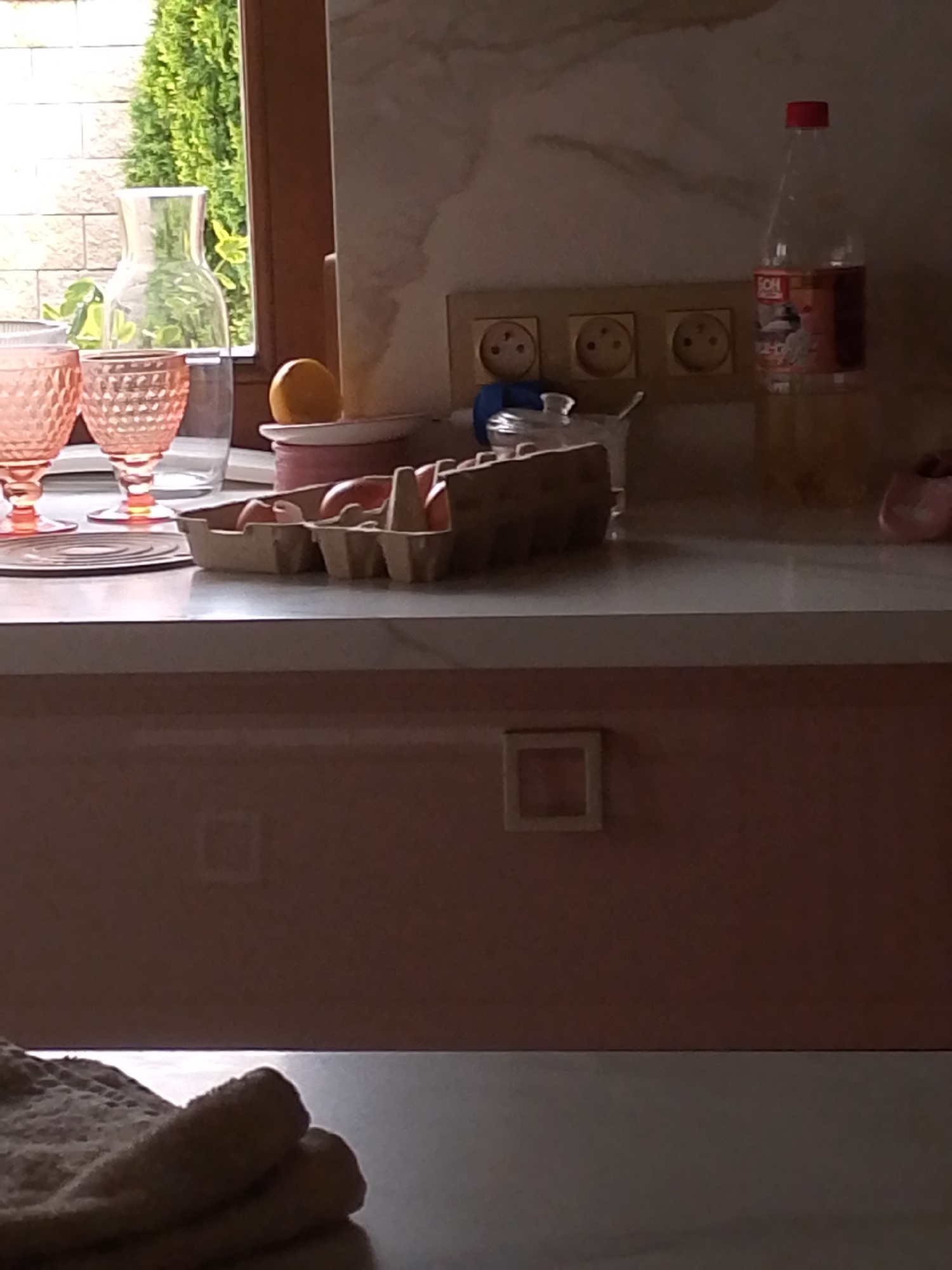 In an indoor setting, possibly a kitchen, a close-up shot focuses on a countertop. To the left, two decorative pink glasses are visible, with a clear glass carafe behind them. A small yellow spherical object, possibly a fruit, rests on a white plate. In the center of the frame, an egg carton holds several eggs. A bottle with a red cap and a label is positioned on the right side of the countertop. Behind the countertop, electrical outlets are set into a light-colored wall. The lower part of the image shows a pink-toned surface with a small square frame on it. The overall lighting suggests it might be daytime, with light entering from a window that is partially visible on the left, illuminating a green tree outside. The scene appears to be static, with no active human presence.