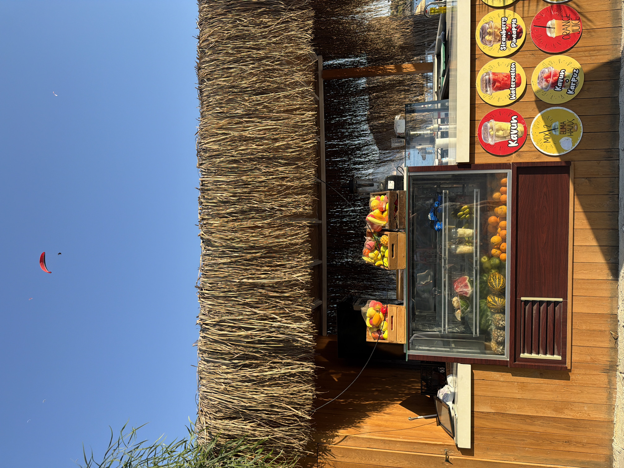 This image depicts a fruit juice stand located on a beach in Fethiye, Turkey. The stand features a thatched roof and wooden walls. In front of the stand, a refrigerated display case showcases various fruits and other items. Several circular signs advertising different juice flavors are mounted on the wooden wall below the counter. In the background, the sea and a beach umbrella are visible, and several paragliders are seen in the clear blue sky. The scene is bathed in warm sunlight, casting long shadows.