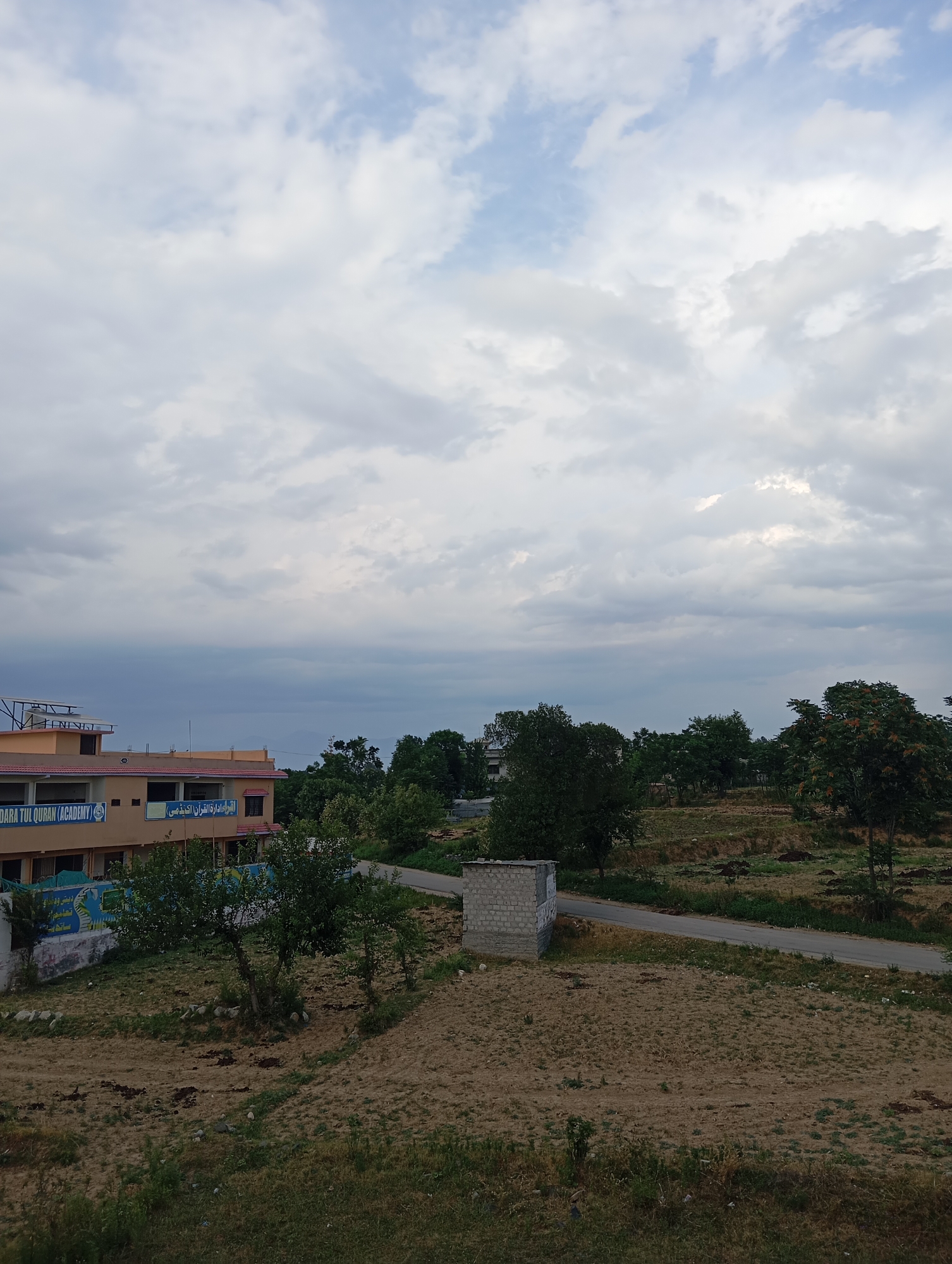 The image captures a view of the Al-Madaris Tul Quran Academy in Dangari, Pakistan, under a cloudy sky. In the foreground, there is a dry, cultivated field with some scattered vegetation. To the left, the academy building is visible, a two-story structure with a tan exterior and red trim. Text in English and Arabic is present on the building's facade. Adjacent to the building are some trees and a blue-and-white mural. A paved road runs through the middle ground, bordered by more trees and grassy, uneven terrain. A small, unfinished brick structure sits to the right of the road. The background consists of rolling hills covered with trees. The overall atmosphere is overcast, suggesting the possibility of rain. There are no people visible in the image.