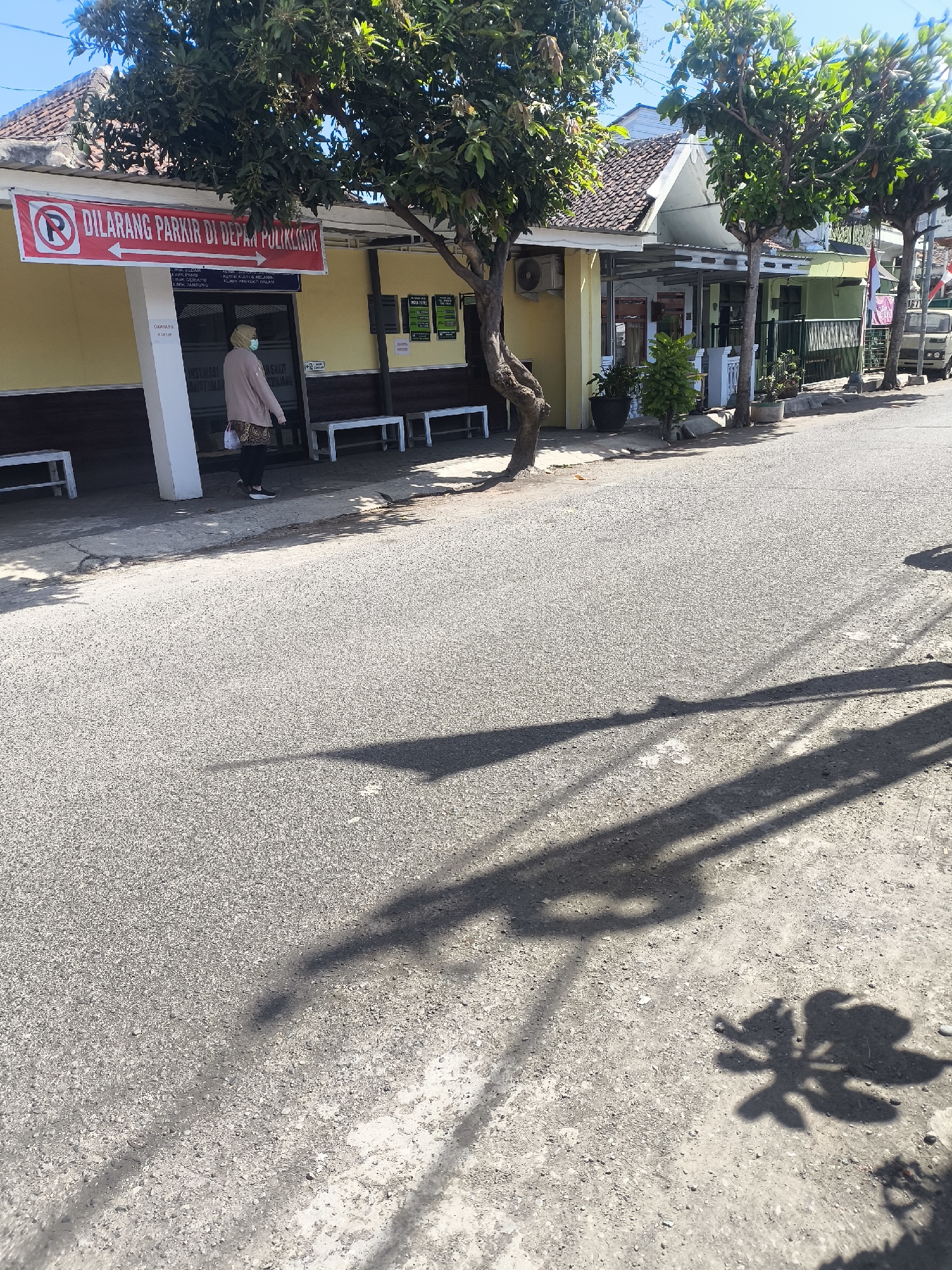 The image depicts a street scene in Lumajang, Indonesia, with a building that appears to be a clinic or polyclinic. A prominent red and white sign with a red circle and slash over the letter "P" indicates "DILARANG PARKIR DI DEPAN POLIKLINIK," meaning "NO PARKING IN FRONT OF THE POLYCLINIC." A woman wearing a light-colored headscarf, face mask, and a light brown jacket is walking away from the entrance of the building. She is carrying a bag over her shoulder. The building has a yellow facade and a tiled roof. Several white benches are placed along the front of the building. Trees line the sidewalk and cast shadows on the asphalt road. The road appears to be in moderate condition with some cracks. The sky is clear and blue, suggesting a sunny day. Shadows of trees and possibly other objects are cast across the road and sidewalk, indicating the direction of sunlight. A white pillar supports the awning of the building. In the background, other buildings and trees are visible, along with a parked vehicle and a small Indonesian flag. The overall impression is a typical daytime scene in a residential or commercial area of the city.