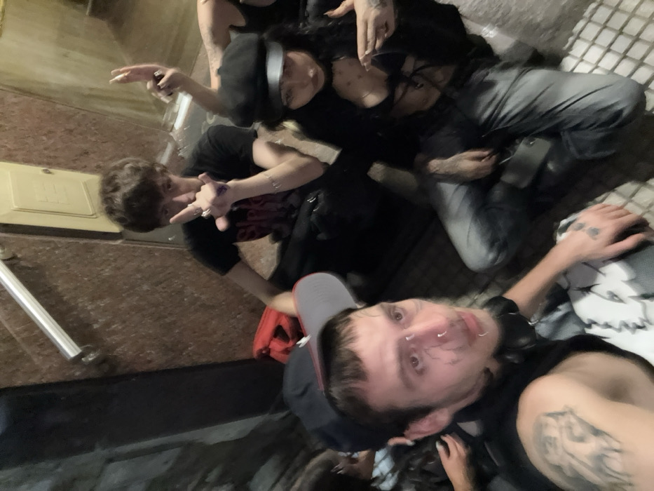 A group of people take a selfie. The foreground shows a man with a backwards baseball cap, facial piercings, and tattoos. In the middle ground, a curly-haired individual gestures with their fingers. Behind them, a person with dark hair and a black beret also gestures. Another person with grey pants is positioned behind. The scene appears to be outside, possibly near a building or structure.