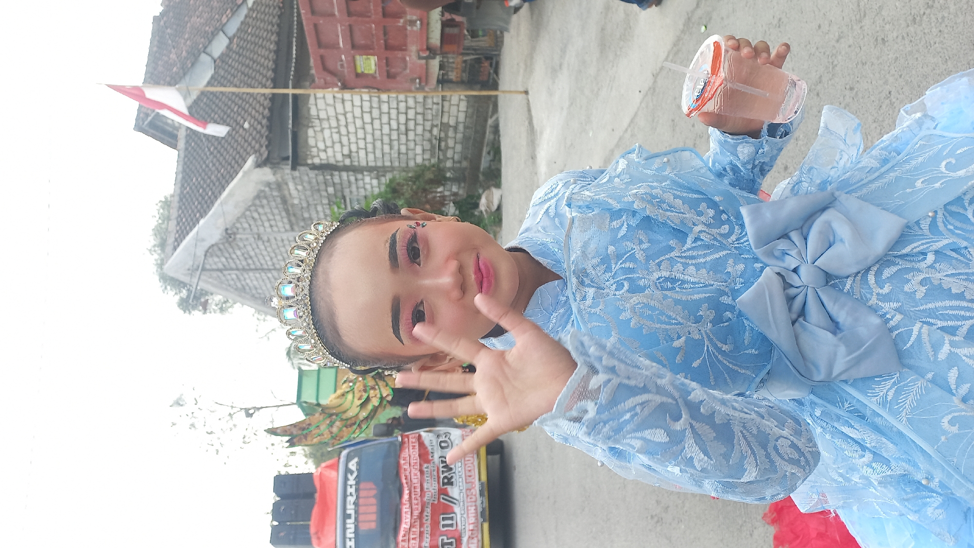 A young girl, adorned with a tiara and elaborate makeup, stands outdoors, raising her left hand. She is wearing a light blue, ornate dress with intricate white embroidery and a large bow at the waist. In her right hand, she holds a clear plastic cup with a pink beverage and a straw. Behind her, a red truck with text that appears to be in Indonesian is partially visible, along with a brick building and a red and white flag on a pole. The ground is paved. The scene suggests a celebration or event in Kedungmegarih, Indonesia.