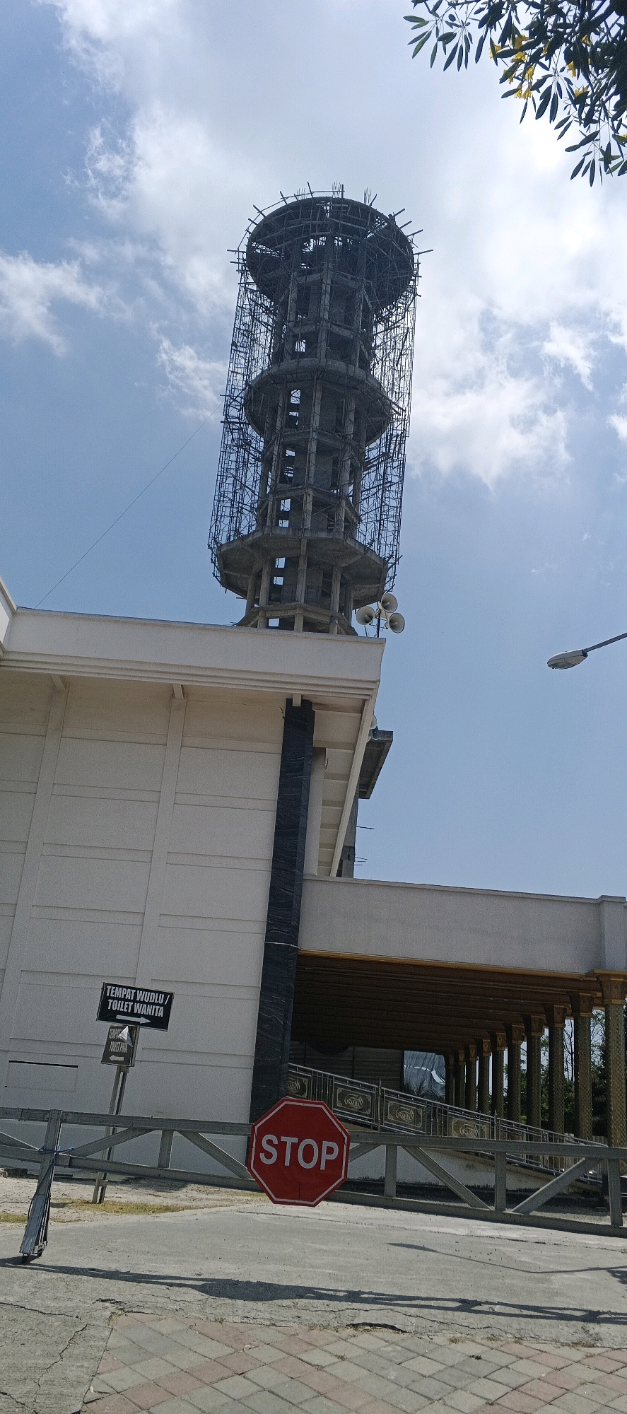 The image shows a construction site in Kandangsemangkon, Indonesia, featuring a partially constructed tower covered in scaffolding. Adjacent to the tower is a white building with a dark vertical accent. A red octagonal stop sign is prominently displayed in the foreground, along with a sign indicating "TEMPAT WUDLU / TOILET WANITA" (Wudlu Place / Women's Toilet). The sky is blue with scattered clouds, and a tree with yellow flowers and leaves is visible in the upper right corner. A street lamp is also present on the right side of the frame. The ground appears to be paved with concrete and interlocking bricks. No people are visible in the image.