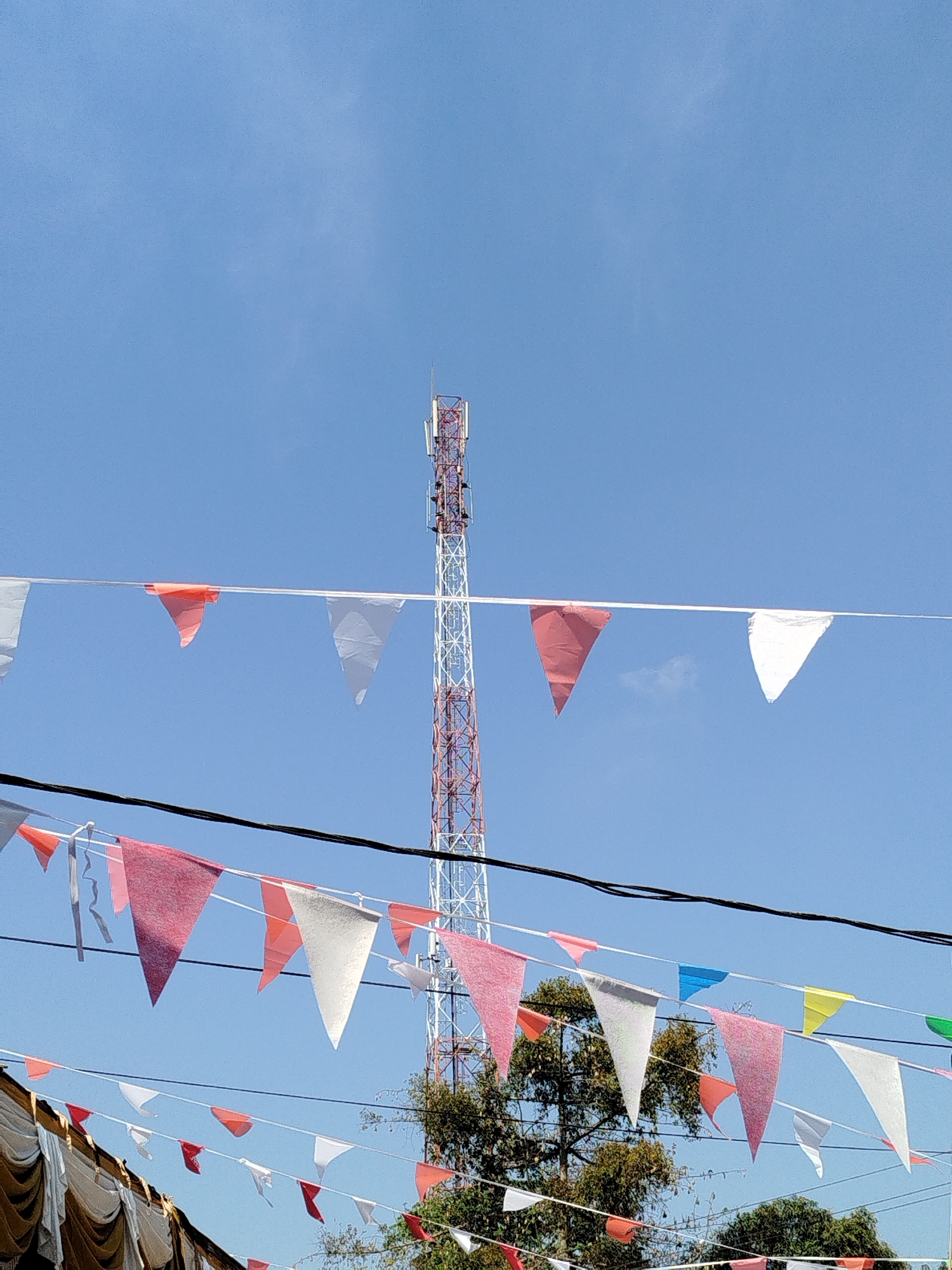 The image captures a clear blue sky above a cellular tower and strings of triangular flags. The flags are a mix of red, white, pink, and yellow, strung across multiple lines. In the foreground, a portion of a tan-colored tent or canopy is visible at the bottom left. The cellular tower, with its red and white lattice structure, stands tall in the background, partially obscured by trees. The scene appears to be outdoors, likely in an open area. The location is Parseh, Indonesia.