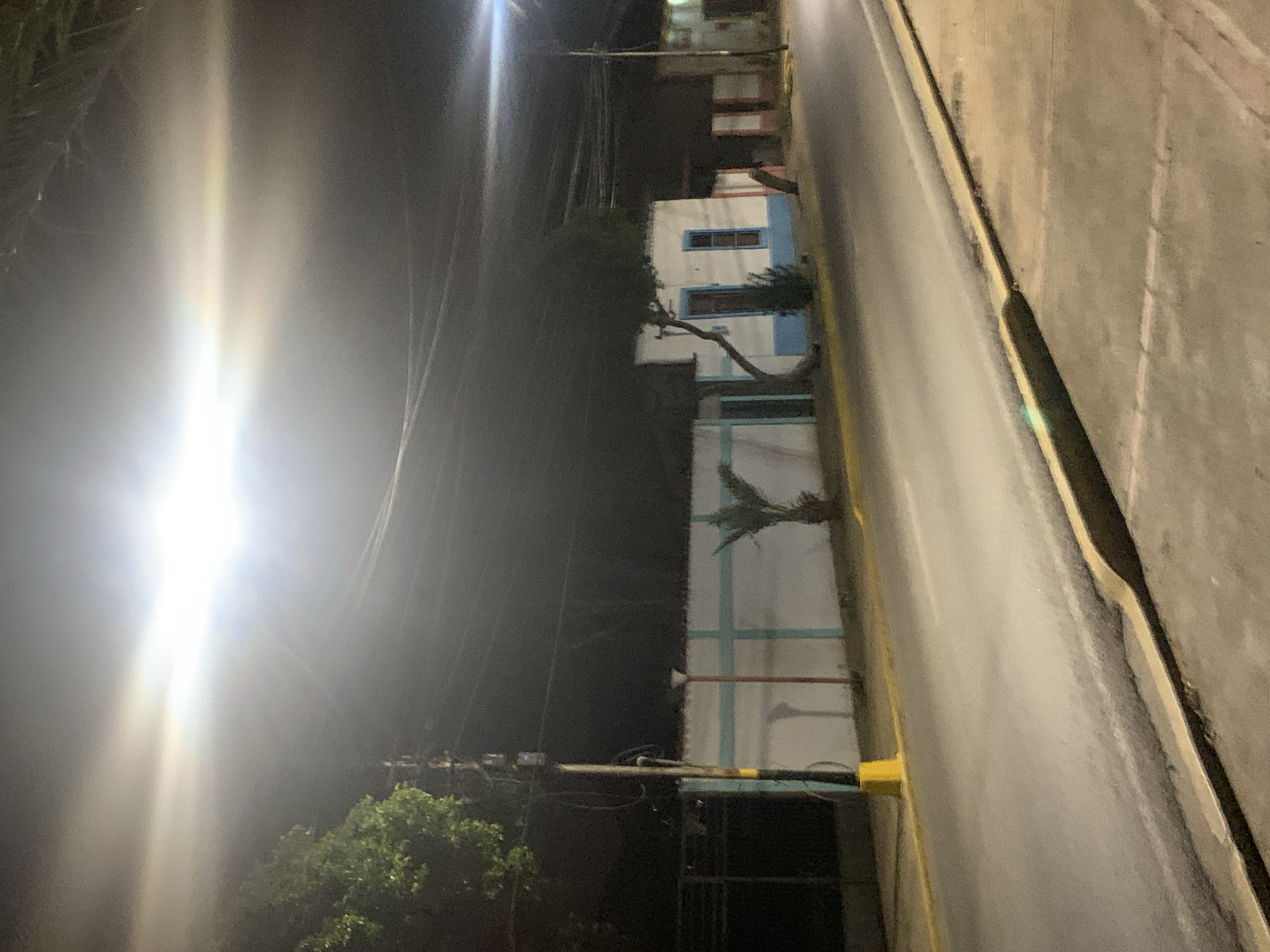 The image depicts a nighttime street scene in Santa Ana, Venezuela. The street is asphalt, with a yellow curb on the right and a yellow and white striped curb on the left. A large, bright light source dominates the upper portion of the image, creating a significant lens flare and overexposing the sky. In the background, a low-rise building with white walls and light blue trim is visible. A tree with a distinctive, gnarled trunk grows in front of the building, leaning over the roof. A small palm tree stands to the right of the building. A utility pole with multiple wires runs along the left side of the street, near the building. The overall atmosphere is dim, illuminated by artificial light.