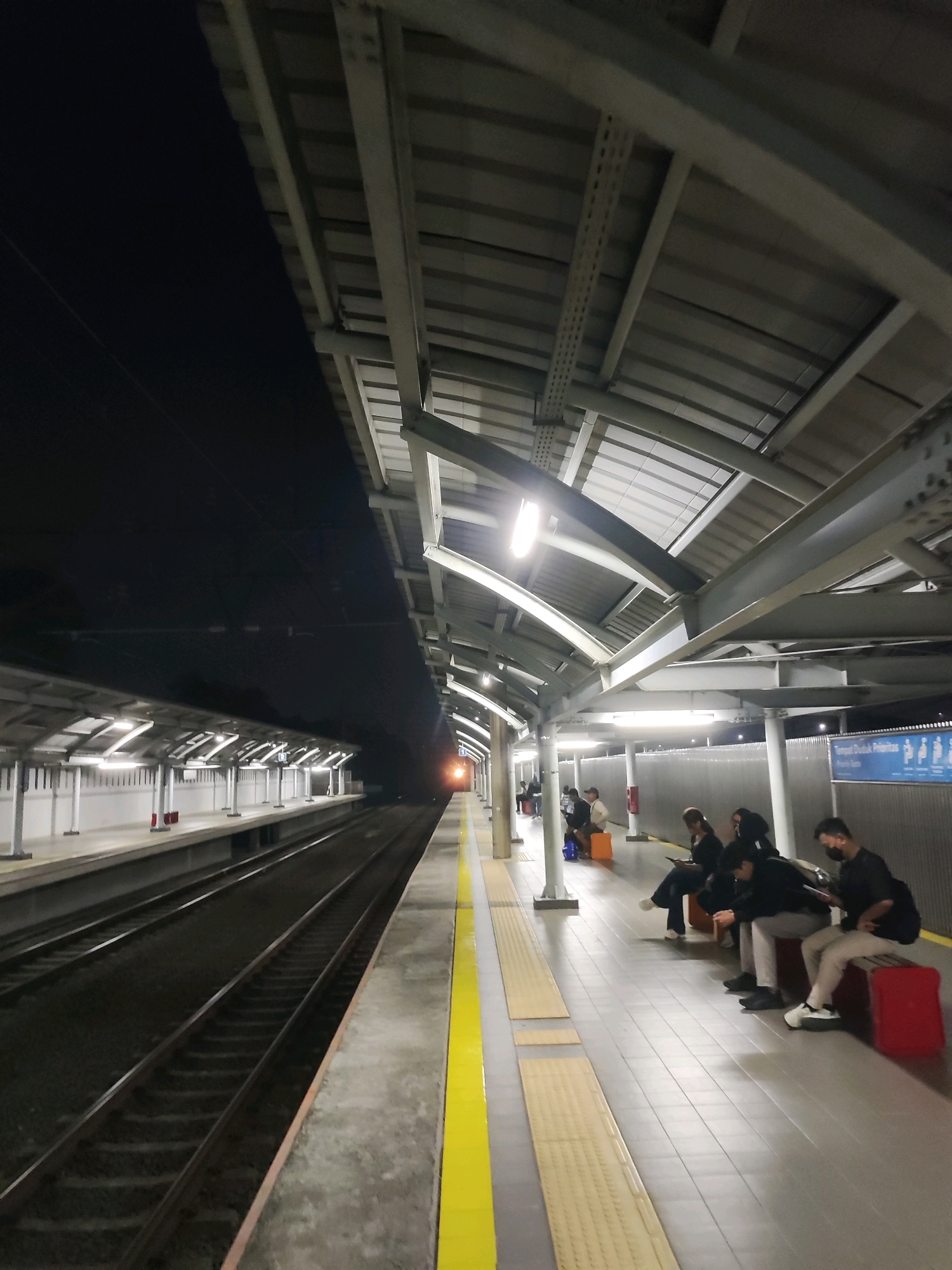 The image depicts a train station platform at night. The platform is covered by a large, modern canopy supported by metal beams. Fluorescent lights illuminate the platform, casting a cool, artificial glow. To the left, the dark expanse of the night sky is visible, with train tracks stretching into the distance. A train, its headlights visible, is approaching from the far end of the platform.
On the right side of the platform, several people are seated on red benches, waiting for the train. Most are wearing casual clothing and some are looking at their phones. The floor of the platform is tiled, with yellow tactile paving indicating the edge of the platform. A blue sign with white text and icons, likely providing information to passengers, is mounted on a fence in the background. The overall atmosphere is one of quiet anticipation.