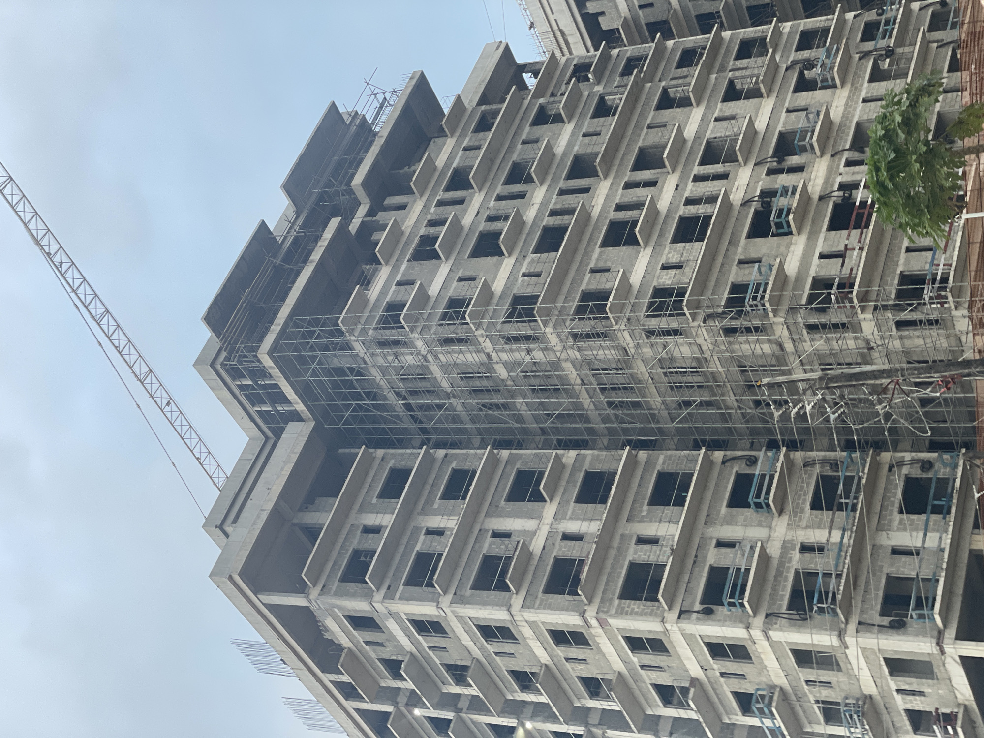 The image shows a tall, multi-story building under construction in Ilado, Nigeria. The structure is made of concrete and appears to be a residential or office complex, with multiple floors featuring balconies and window openings. Scaffolding is visible on the exterior of the building, indicating ongoing work. A large construction crane is positioned above and to the right of the building, with its boom extending across the sky. The sky is overcast and appears to be a light gray color. A small green tree is partially visible in the lower right corner of the image. The scene depicts a typical urban construction site in Nigeria.