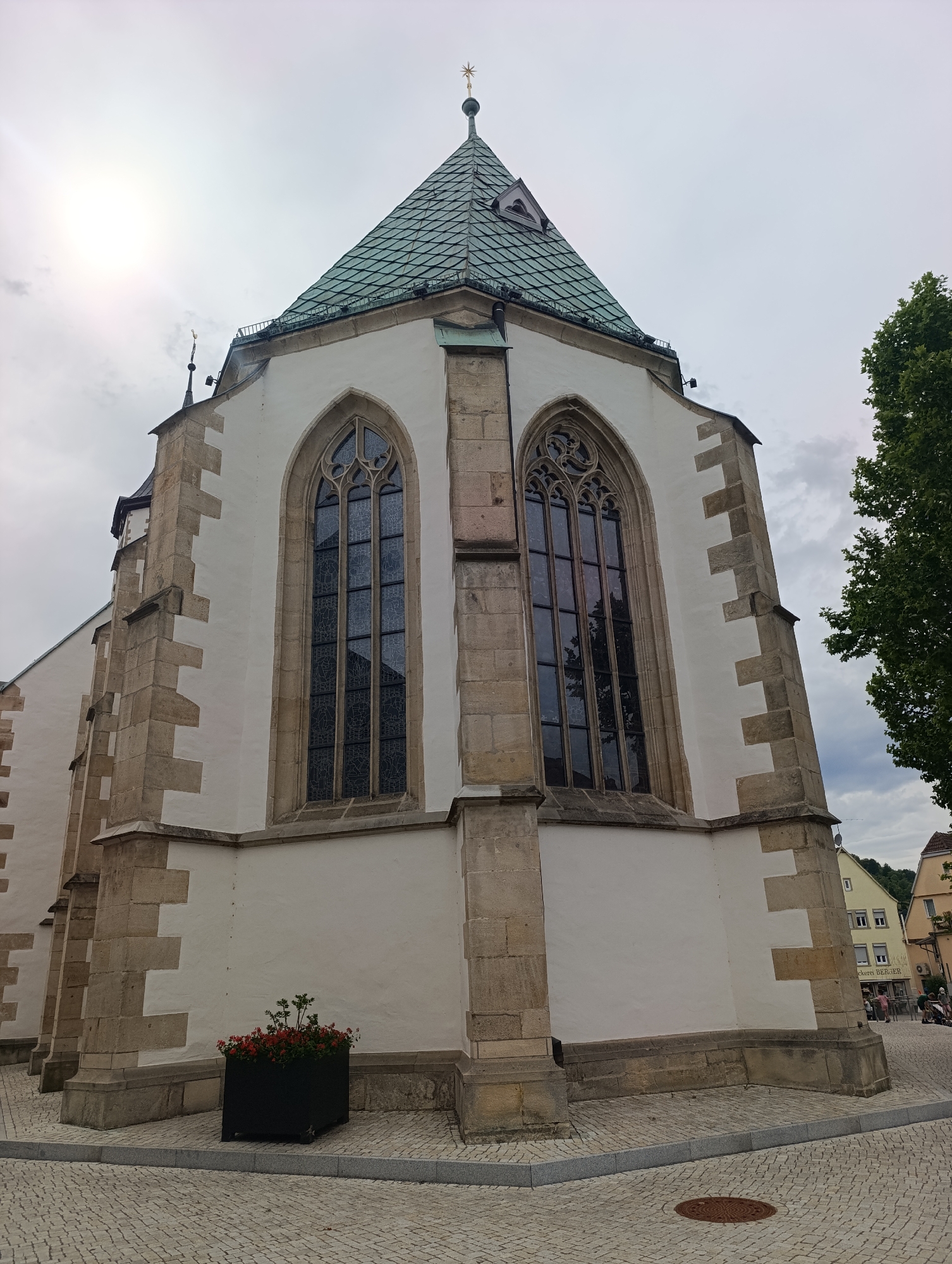 The image shows the exterior of the historic St. Michael's Church in Pfullingen, Germany. The church has a distinctive white facade with rough-hewn stone accents at the corners and around the Gothic-style windows. The roof is green and steeply pitched, topped with a spire. In the foreground, the church is surrounded by a cobblestone plaza. To the right of the church, a large green tree is visible, and beyond that, a street with buildings and a few people. The sky is overcast, suggesting it may be a cloudy day. The overall impression is one of historical architecture and a typical town square setting.