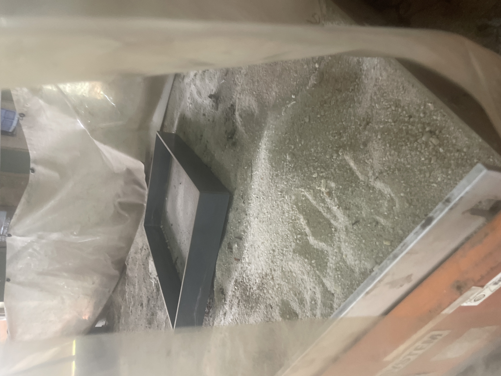 The image shows a shallow, dark rectangular frame resting on a bed of light-colored gravel and sand. The frame appears to be made of metal and is empty. The surrounding area is covered with the same gravel and sand, with some areas appearing more compacted than others. In the background, a translucent white tarp is draped, obscuring a portion of the scene and creating a soft, diffused light. The tarp is attached to an unseen structure, as evidenced by visible grommets and possible attachment points along its top edge. To the left of the frame, a portion of an orange and metallic object, possibly equipment or machinery, is visible, with faint lettering on its surface. The overall environment suggests an outdoor or partially enclosed construction or work site. No individuals are present in the frame, and no specific actions are depicted. The location in Ihiagwa, Nigeria is not visually indicated in the image itself.