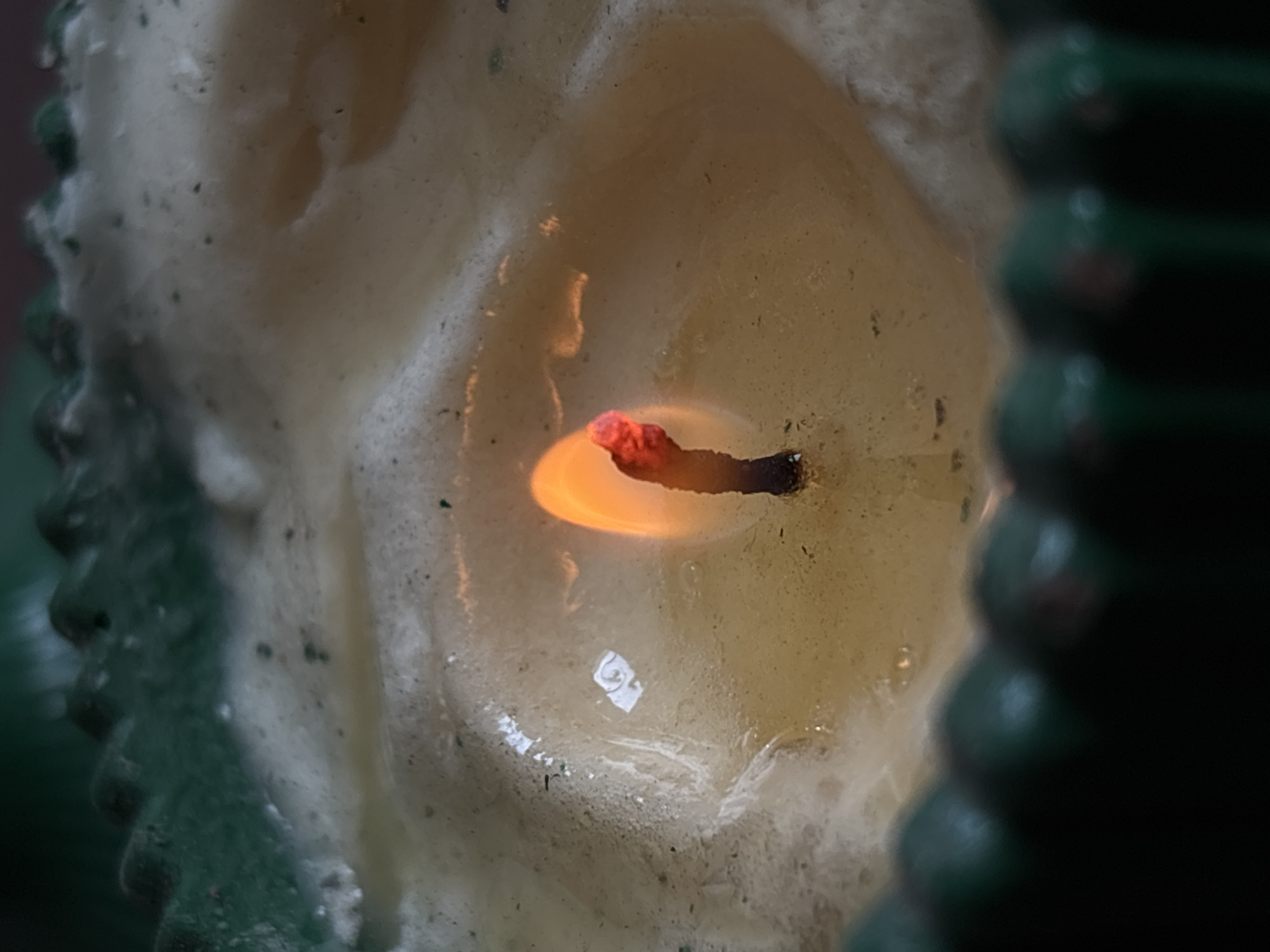 A close-up shot displays the burning wick of a candle, nestled within a pool of molten wax. The candle itself seems to be green, with a textured outer surface. The interior of the candle appears to be a light yellow color with some dark spots. There is no indication that the photo was taken in Santa Clara Ocoyucan, Mexico. There are no people in the photo.