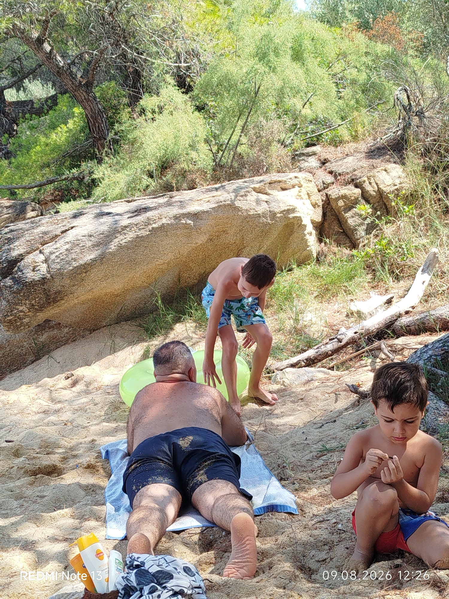 The image depicts a sandy beach scene with natural rocky and vegetated surroundings, likely within the Sithonia Municipal Unit, Greece, given the context. In the foreground, an adult male is lying on his back on a beach towel, facing away from the camera. He is wearing dark swim trunks. To his right, a young boy is kneeling on the sand, looking down at something in his hands. He is wearing red and blue swim trunks. Further back, another young boy, facing away from the camera, is standing and looking down. He is wearing patterned swim trunks and is near a large, light-colored inflatable ring. Behind the figures, large rocks and dense green vegetation are visible, with sunlight filtering through the trees. The overall atmosphere is one of leisure and outdoor activity.