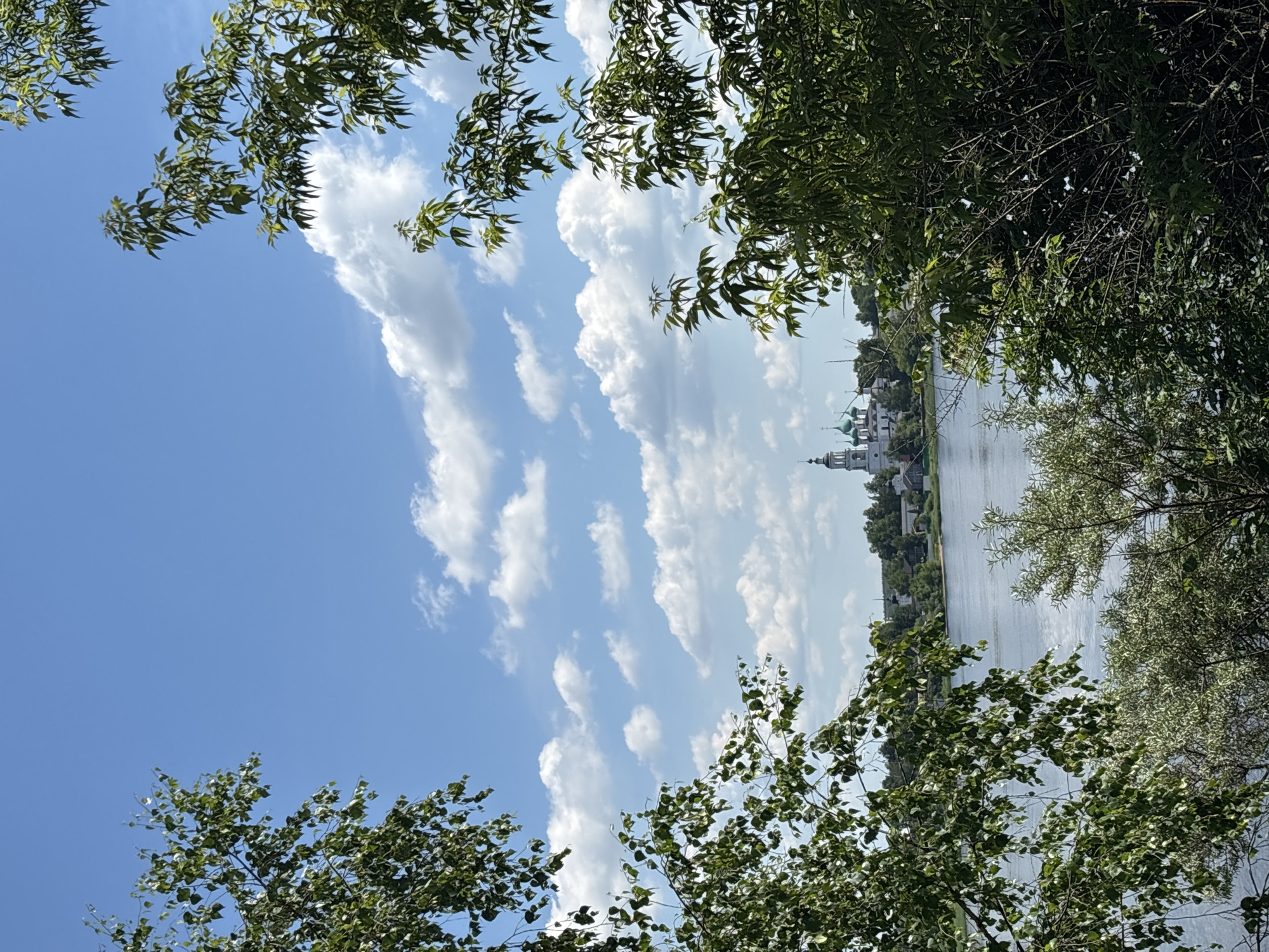 The image captures a serene natural landscape with a body of water in the foreground and a distant town with a prominent church. The scene is framed by lush green trees on either side, their leaves creating a natural border. The sky above is a vibrant blue, dotted with fluffy white clouds, suggesting fair weather.
The water, appearing calm and reflecting the sky, stretches across the middle ground. On the far bank, a settlement is visible, characterized by buildings with traditional architecture. Dominating the skyline of the settlement is a white church with several green domes, a common feature in Russian Orthodox churches. The church's spire and architectural details are distinct, suggesting it is a significant landmark.
The foreground is dominated by dense foliage from trees and bushes, creating a sense of depth and natural enclosure. The branches and leaves of these trees are in sharp focus, contrasting with the softer, more distant view of the town and water. The overall impression is one of natural beauty and tranquility, with a touch of historical or cultural significance provided by the church. The lighting suggests it is daytime, likely late morning or early afternoon, given the bright sky and clear visibility. The environment appears to be a rural or semi-rural setting, possibly a park or a natural preserve adjacent to a body of water and a settlement.